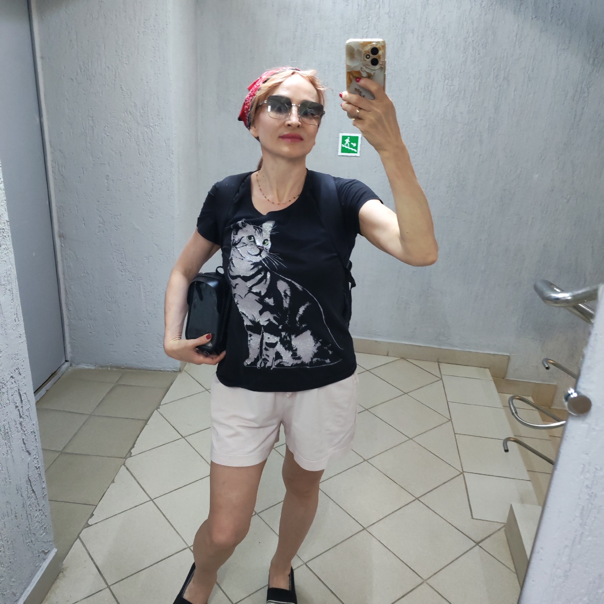 A woman is taking a selfie in a hallway with beige tiled floors and textured, light gray walls. She is wearing a red bandana, sunglasses, a black t-shirt with a cat print, light pink shorts, and black shoes. She has a black backpack on her shoulders and is holding a small black bag in her left hand. An exit sign is visible on the wall behind her. The reflection of a staircase with a metal handrail is visible in a mirror to her right. This is an interior shot. Based on previous knowledge of the user, the location is likely the city of Chaikovsky, Russia.