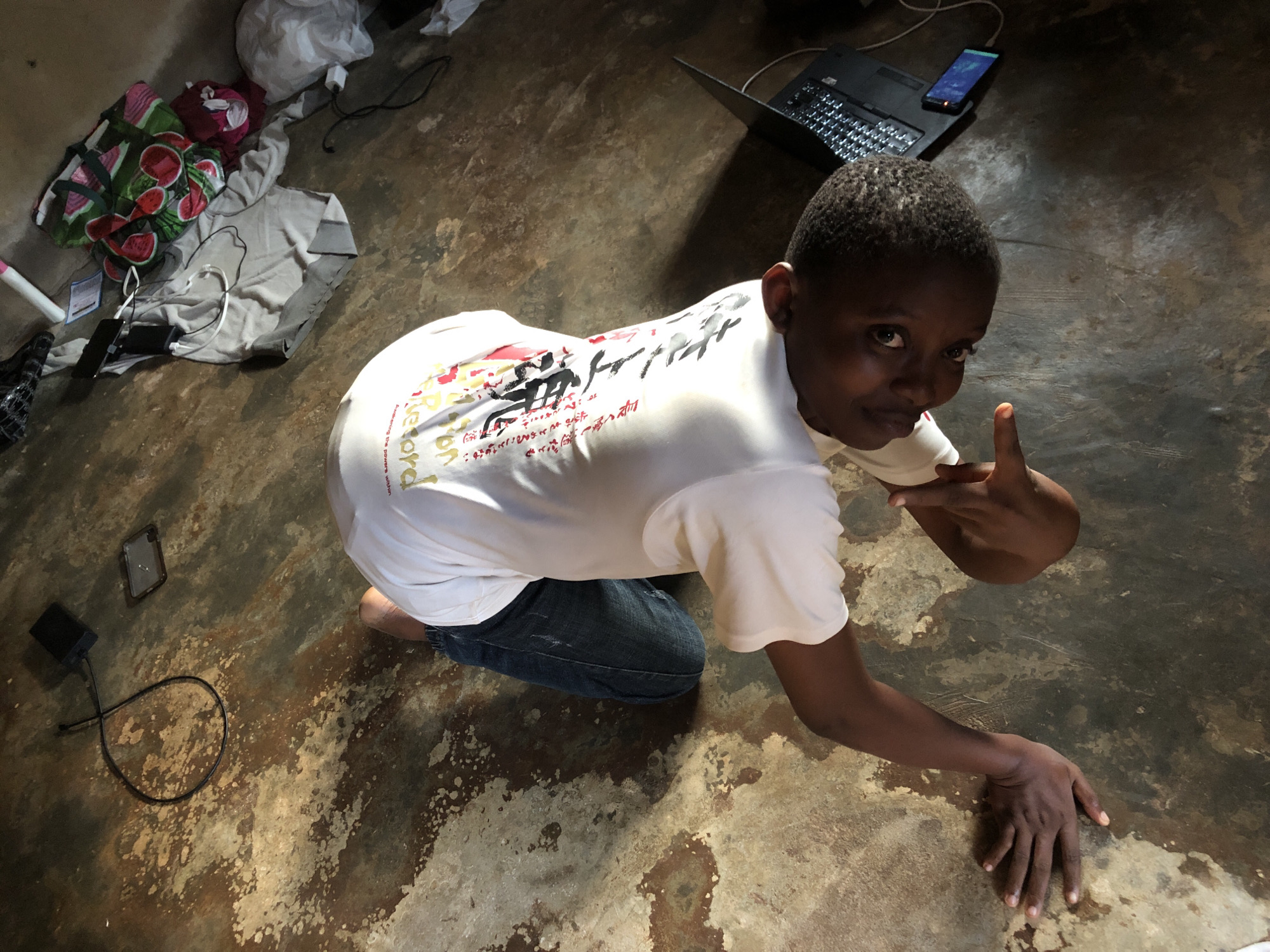 A person is kneeling on a mottled brown and tan floor. They are wearing a white t-shirt with graphics and dark denim jeans. To the left is a pile of clothes and accessories. A laptop and smartphone are at the top right of the frame. The location is the city of Unknown, Unknown. The person is gesturing with their hand, giving a thumbs up.
