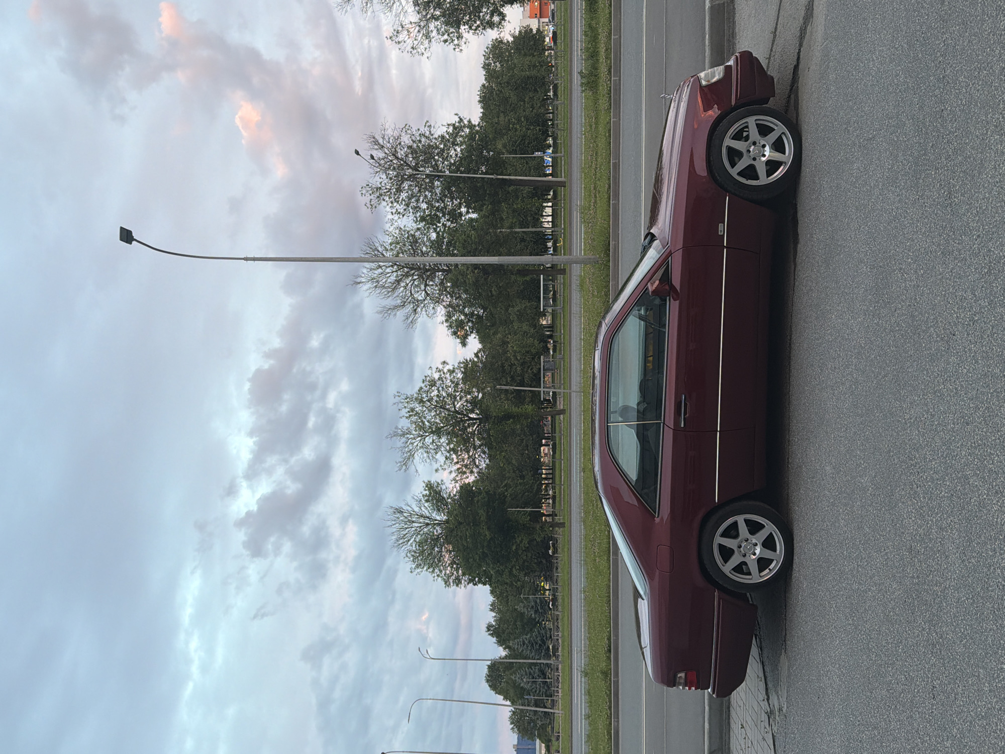 A burgundy two-door coupe, likely a Mercedes-Benz E-Class coupe from the 1990s, is parked on the side of a road. The car is facing to the left of the frame, with its side profile visible. It has silver multi-spoke rims. The car is parked on an asphalt surface adjacent to a sidewalk. Beyond the sidewalk is a grassy median strip, followed by a multi-lane road with some traffic visible in the distance. A row of trees with dense green foliage lines the far side of the road, with a cloudy sky overhead that has hints of pink and orange, suggesting it might be dusk or dawn. Several streetlights are visible, including one tall, curved pole directly behind the car. The background also includes some buildings and what appear to be bus stops. The overall scene depicts a parked vehicle in an urban or suburban environment with natural elements.