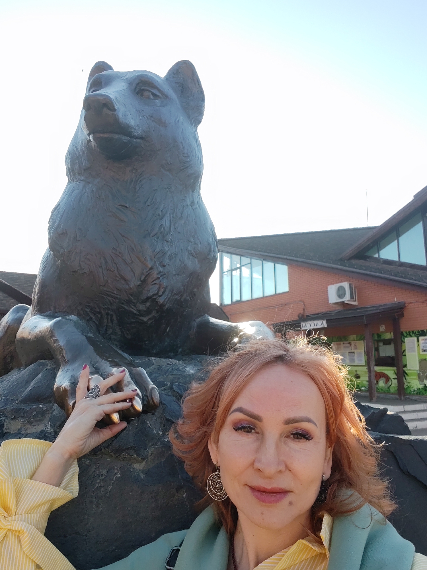 A woman is taking a selfie with a large, dark bear statue. The statue depicts a seated bear with a thoughtful expression. The woman is wearing a yellow striped shirt, a green jacket, and decorative earrings. She has reddish-blonde hair and is smiling slightly. She is touching the bear's paw with her left hand, which has a large ring on it. The background shows a red brick building with windows and an air conditioning unit, a wooden porch with posters, and some greenery. The scene is likely taken in Izhevsk, Russia, given the bear statue is one of the city's most recognizable symbols.
