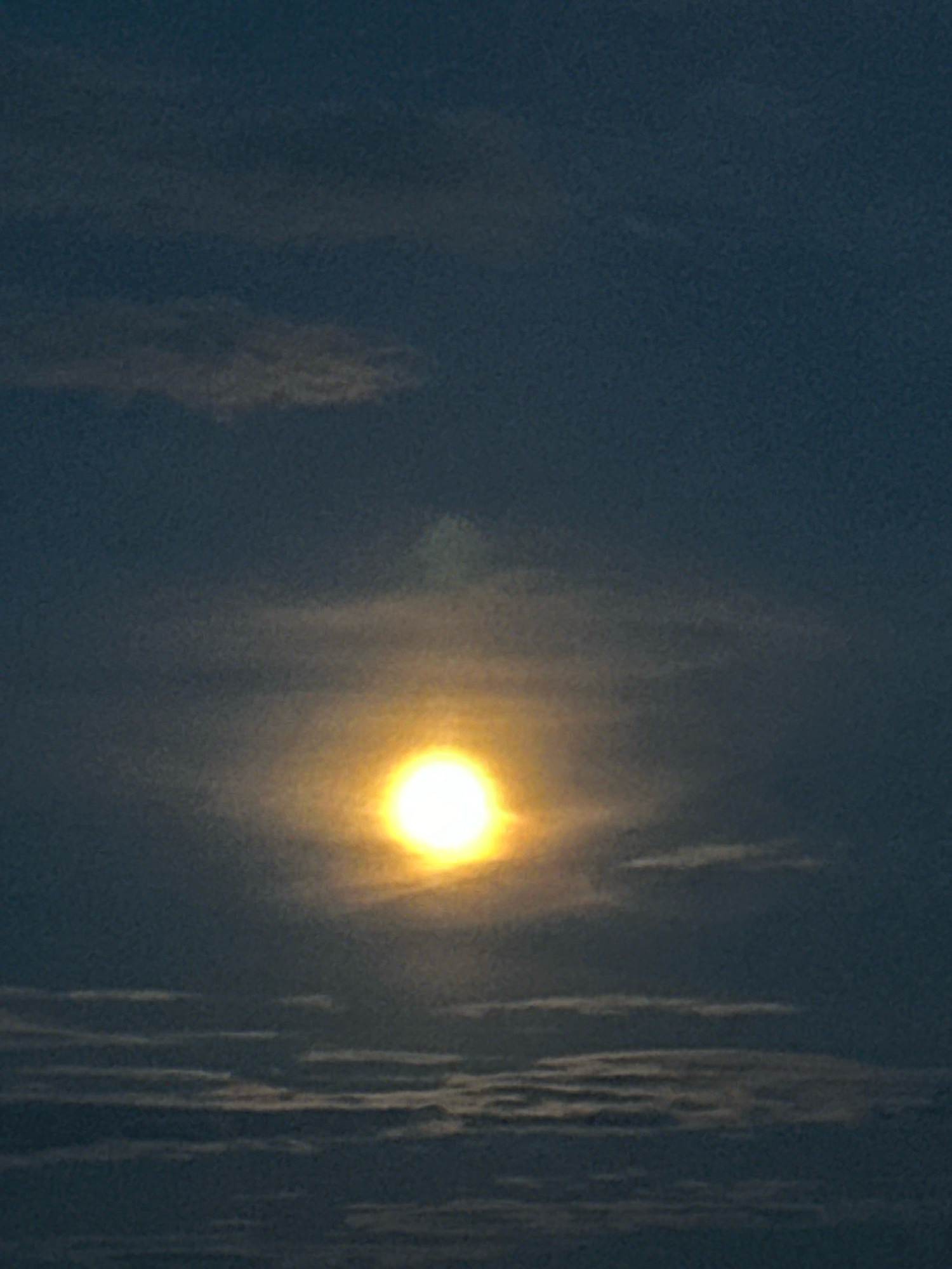 A wide shot captures the sun setting over a flat, dark landscape, possibly in Нарацкі сельскі Савет, Belarus. The sun, a bright yellow orb, is positioned slightly left of the center of the frame, its light diffused by thin clouds that create a hazy halo around it. These clouds appear as wispy streaks across the darker blue sky, with more defined, horizontal cloud formations near the bottom of the image. The overall impression is of a tranquil, natural scene at twilight.