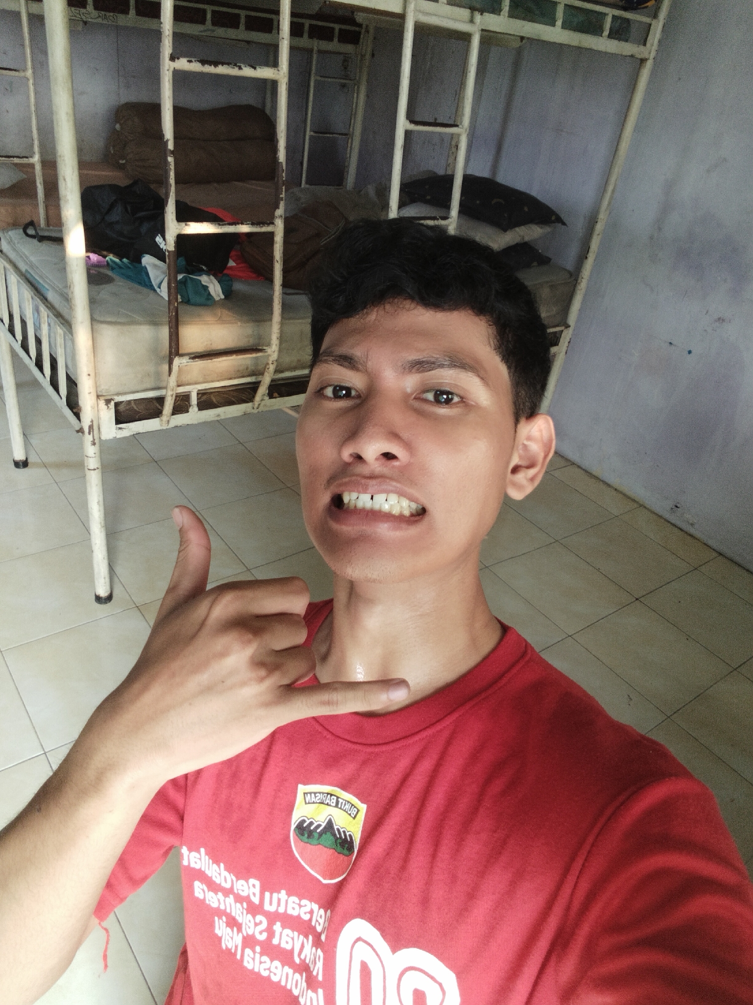 A young adult male with dark hair and medium skin tone is positioned in the foreground, taking a selfie. He is wearing a red t-shirt with a circular emblem featuring mountains and trees. His right hand is raised, forming a "shaka" gesture. Behind him, a metal bunk bed frame is visible, with mattresses and bedding on the lower bunk. The floor is tiled. The lighting suggests an indoor environment, possibly a dormitory or shared living space. The overall scene is casual and appears to be set in a simple living quarter.