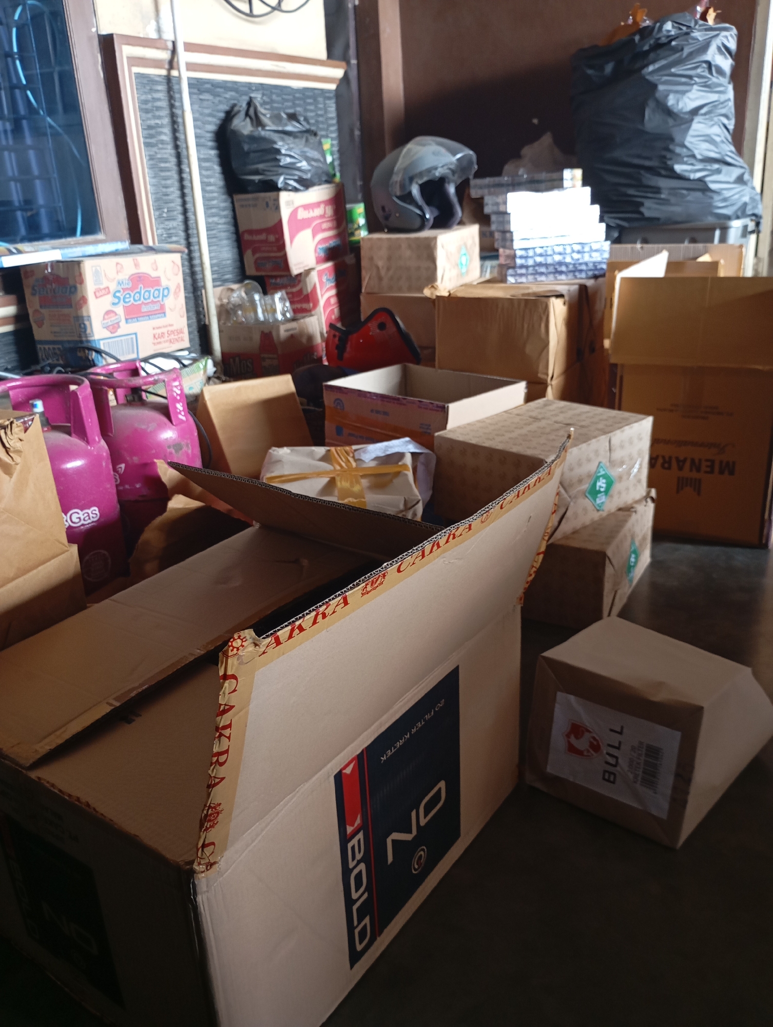 The image depicts a cluttered storage area with various boxes and items. In the foreground, there is a large open cardboard box with "AKRA BOLD" printed on its side, and further to the right, a smaller cardboard box with a "BULL" label. In the background, there are several stacks of boxes, some with visible branding like "Sedaap". Two pink gas cylinders are prominent on the left side. A dark plastic bag is piled on top of some boxes. A motorcycle helmet is visible on a box. The environment appears to be indoors, possibly a warehouse or a storage room, with a concrete floor. The lighting suggests it is daytime. The location is identified as a storage area in the city of Metro, Indonesia.
