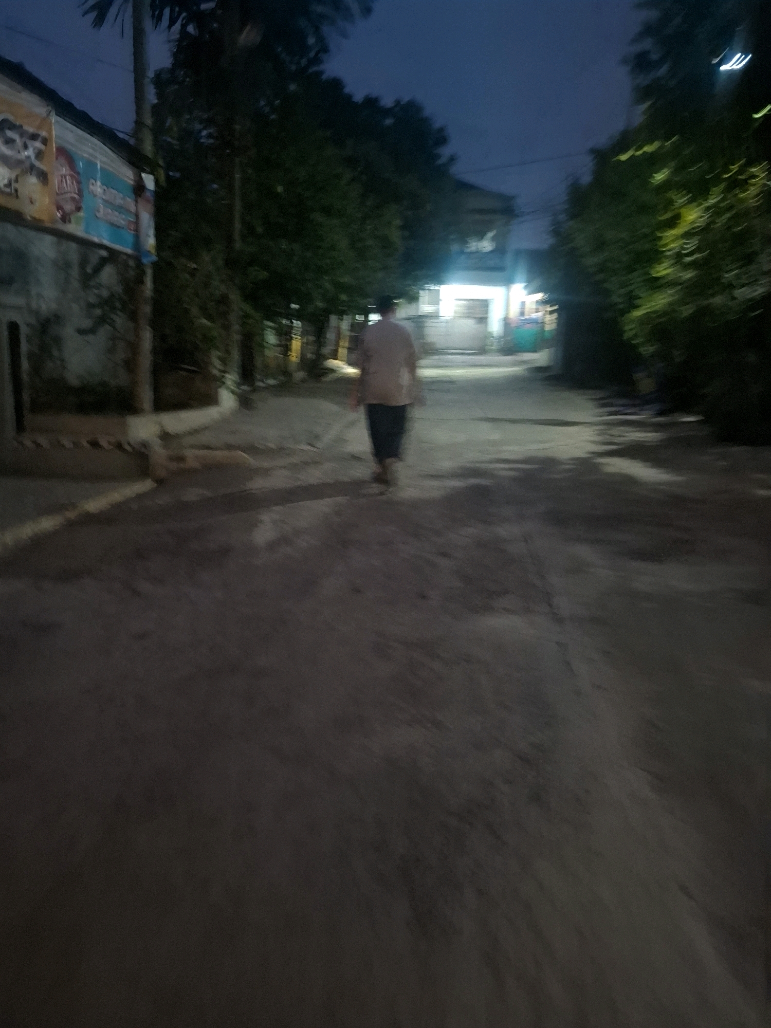 The image depicts a dimly lit street scene, likely in a residential area, at dusk or night. A single person, appearing to be an adult, is walking away from the camera down a paved, somewhat uneven road. They are wearing a light-colored shirt and dark pants. The environment features trees lining the sides of the road, and the background shows the illuminated facade of a building, suggesting a residential or small commercial area. Signs are visible on a wall to the left, partially obscured and unreadable due to the low light and blurriness. The overall impression is one of a quiet, ordinary street in Indonesia, possibly in the city of Ciheulang Tonggoh, given the context.