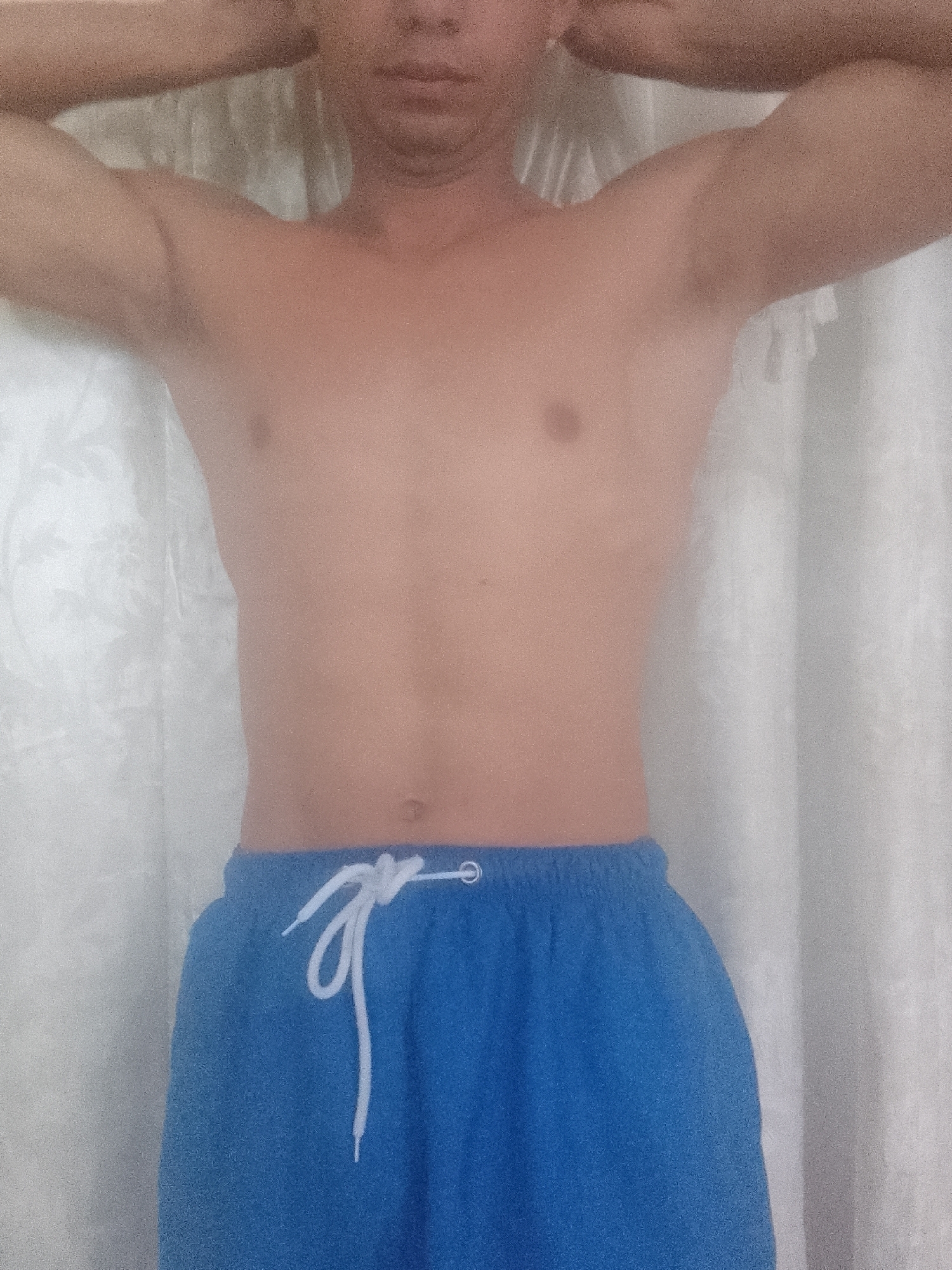 A person with a light complexion stands indoors against a white curtain. The person is shirtless and wearing blue shorts with a white drawstring. The person's arms are raised above their head. The location of Juan Griego, Venezuela, is not visually evident in the image.