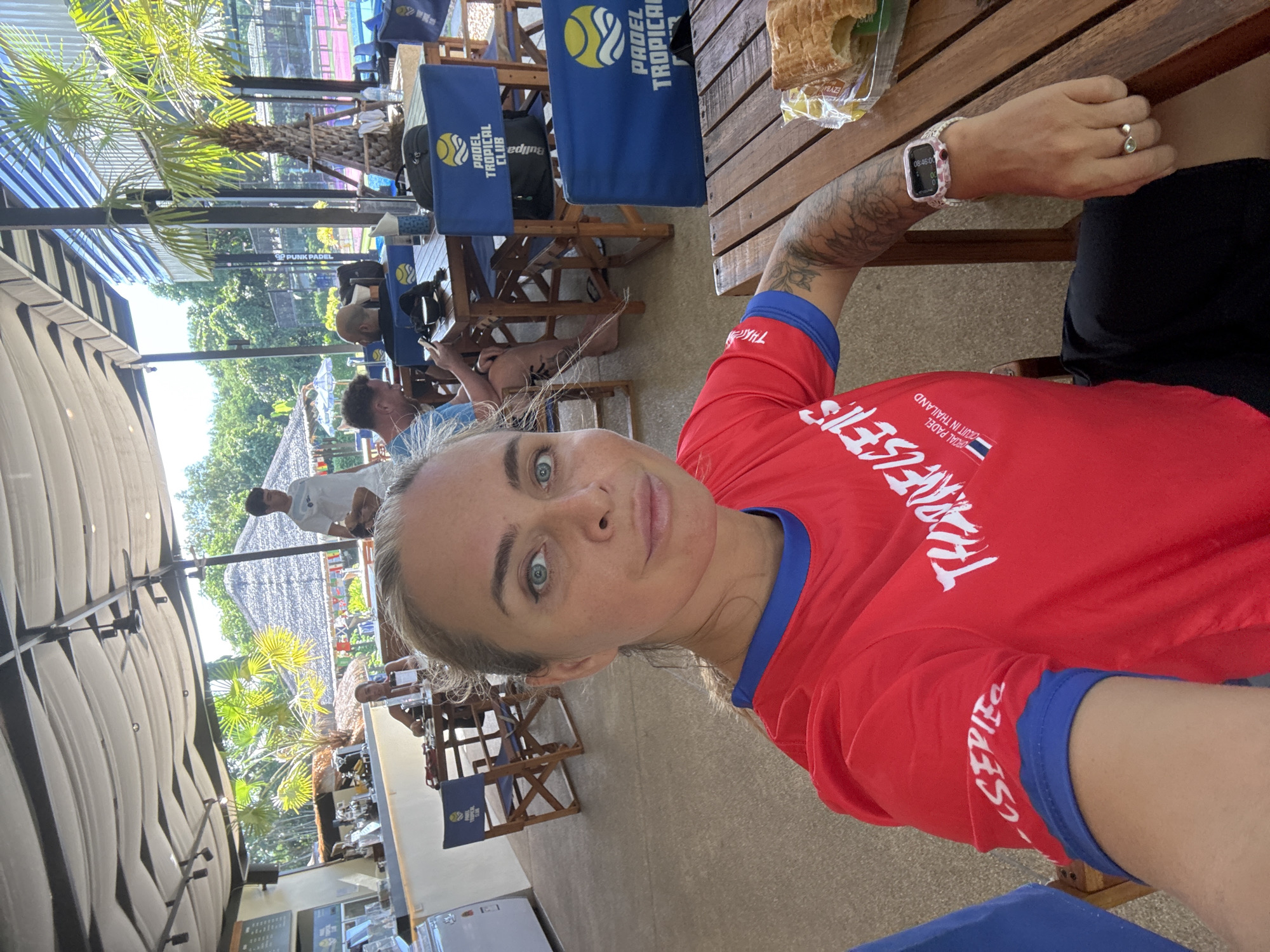 A woman with blonde hair and blue eyes takes a selfie in an outdoor cafe. She is wearing a red t-shirt with blue trim, and a tattoo is visible on her arm. She is sitting at a wooden table, and a pastry is on a plate next to her. In the background, other people are seated at tables, and palm trees are visible. The cafe appears to be part of a Padel Tropical Club, indicated by the blue chairs with the club's logo. The location is identified as Ban Bang Rak, Thailand.