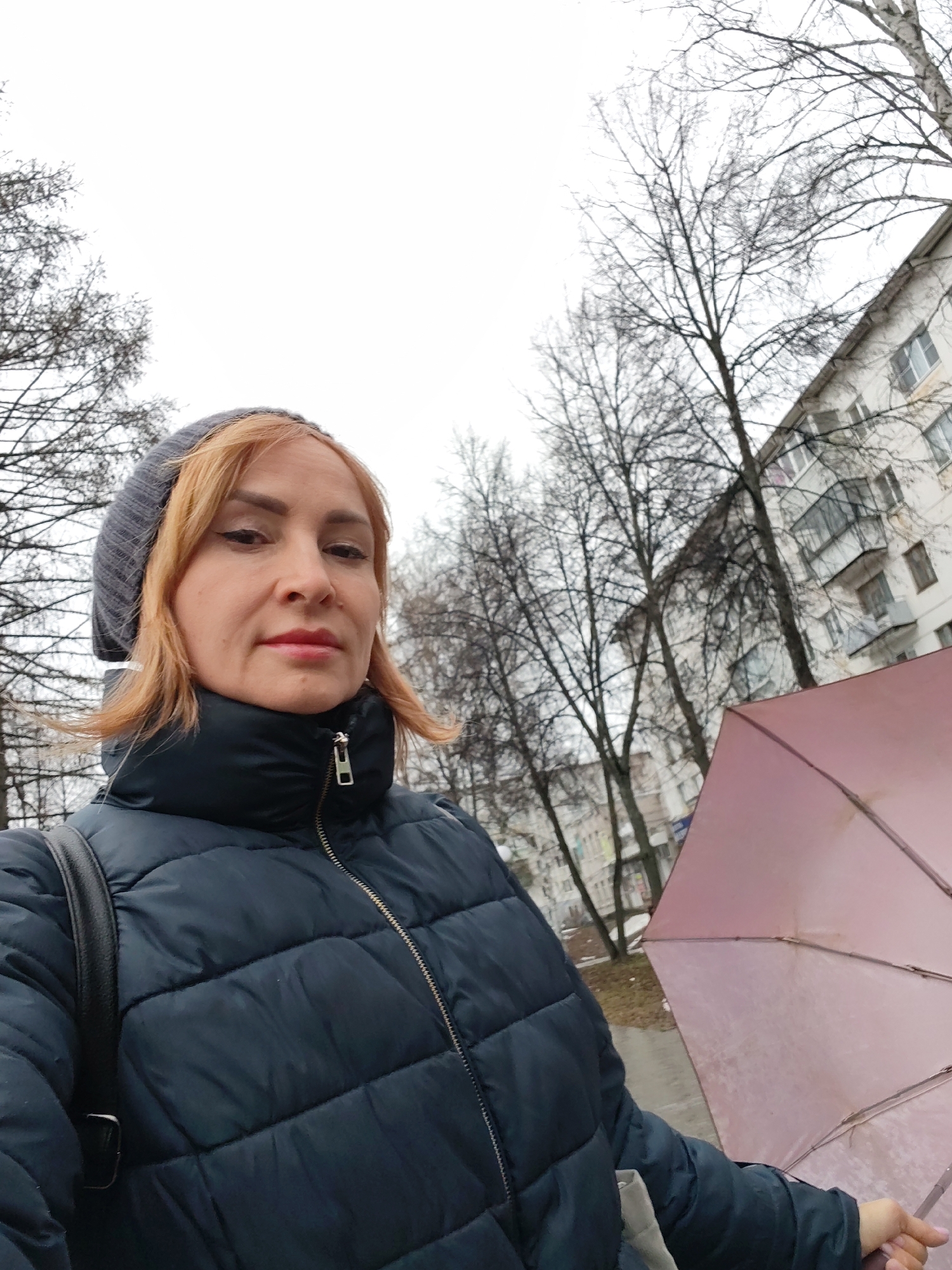 A woman is taking a selfie outdoors on an overcast day. She is wearing a dark blue puffy jacket, a gray knitted beanie, and carrying a black shoulder bag. She is holding a closed, light pink umbrella. Behind her are bare trees, an apartment building, and what appears to be a street. While it is difficult to confirm the exact location in Chaikovsky, Russia, the architecture and general environment suggest a typical urban residential area in a Russian city.