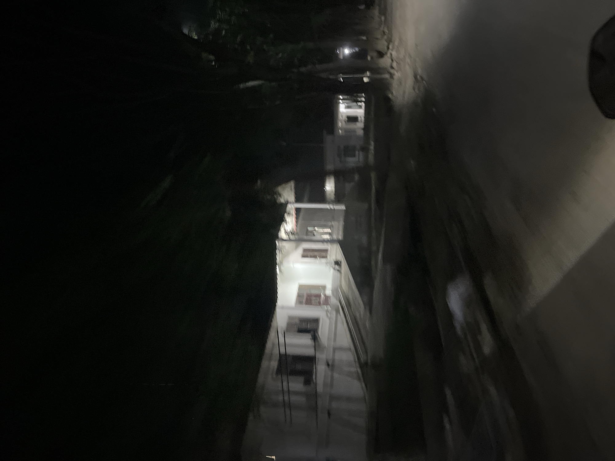 The image shows a night scene in the city of Bundar, Indonesia. A white, low-rise building with multiple windows is visible on the left side of the frame. The building appears to be illuminated from within, casting light onto the surrounding area. A dark road or pathway is in the foreground, and trees with dark foliage line the right side of the road. The overall impression is one of a quiet, possibly residential or institutional area at night. The lighting is dim, and the image has a slightly blurry quality, suggesting movement during capture. There are no people clearly visible in the image.