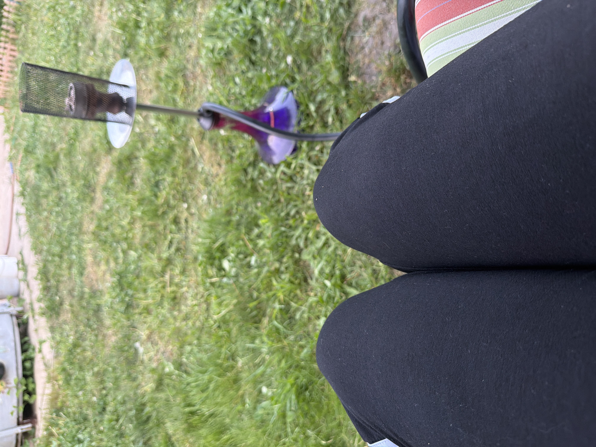 The image shows a hookah on a grassy lawn next to a seated person. The hookah has a purple glass base and a metal and mesh bowl. The person is wearing black leggings and sitting on a chair with striped cushions. The scene appears to be outdoors. The location is Unknown, Unknown.