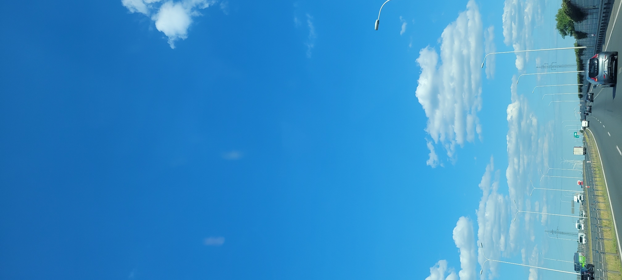The image depicts a bright, clear day with a vivid blue sky filled with scattered white clouds. The perspective is from a moving vehicle, likely on a highway, as indicated by the road, lane markings, and a line of vehicles stretching into the distance. Streetlights line the side of the road at regular intervals. The right side of the image shows a highway with several cars and trucks traveling in the same direction. A gray car is prominent in the foreground on the right. The scene suggests a daytime journey, possibly during summer or a clear spring day given the bright sunlight and the nature of the clouds. The overall impression is one of travel and open road under a beautiful sky. There is no visible text in the image.