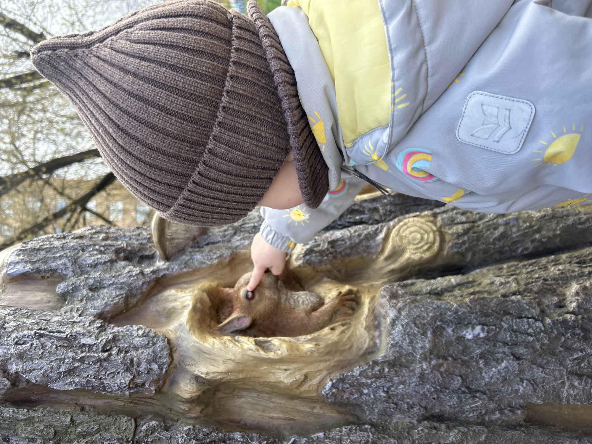 A young child wearing a brown knitted cap and a gray jacket with a yellow lining and small rainbow and sun patterns is reaching out with their right index finger towards a carving of a brown rabbit nestled inside a hollowed-out section of a faux-wood or concrete structure. The structure resembles a section of a log with textured bark and simulated wood grain. The location is Klin, Russia.