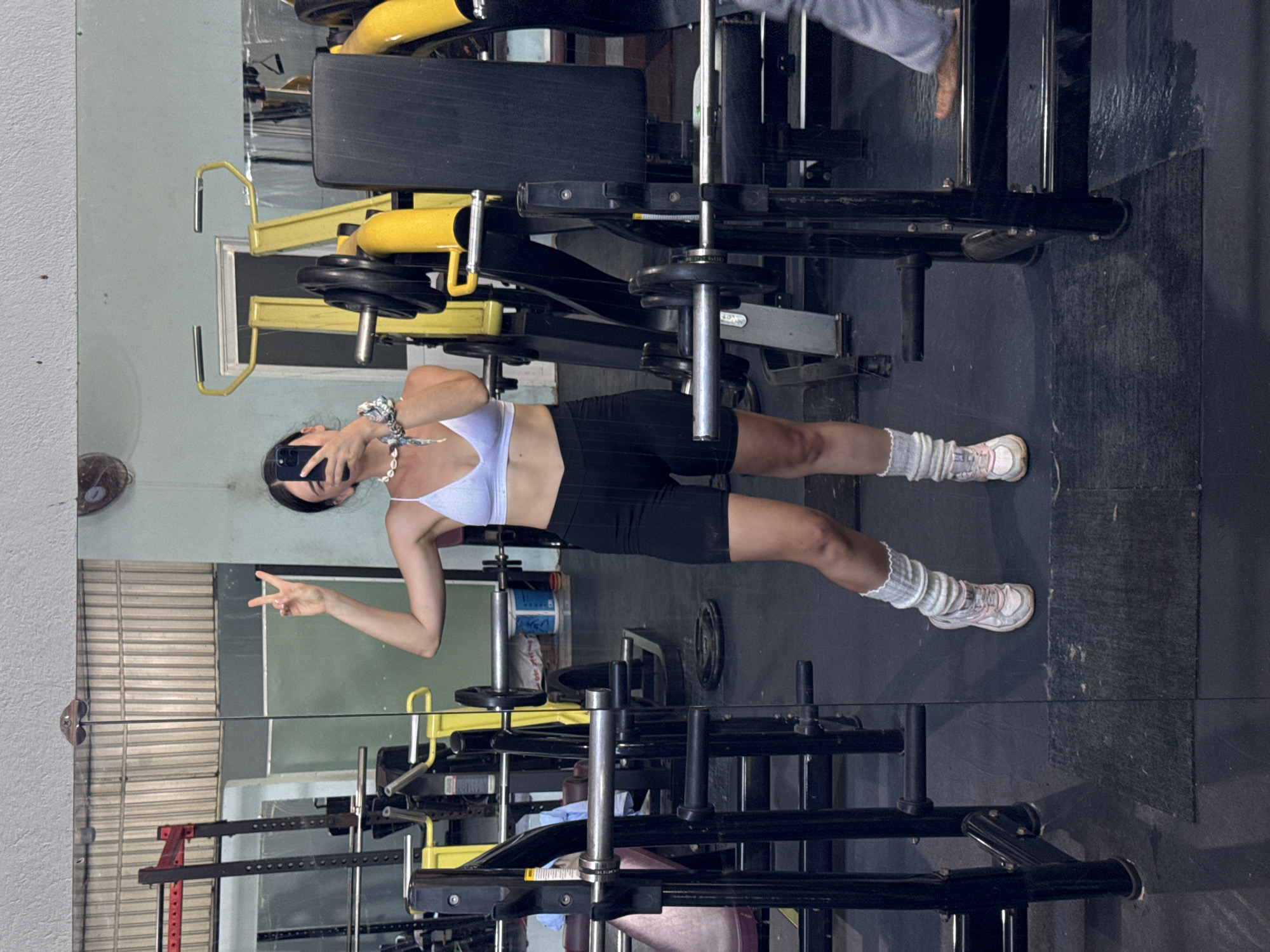 A person stands in a gym in Mũi Né, Vietnam, reflected in a mirror. The person wears a white sports bra, black shorts, white leg warmers, and sneakers, holding a cell phone and making a peace sign. Gym equipment including weight machines and free weights are visible around them. The gym has a black floor and pale green walls.