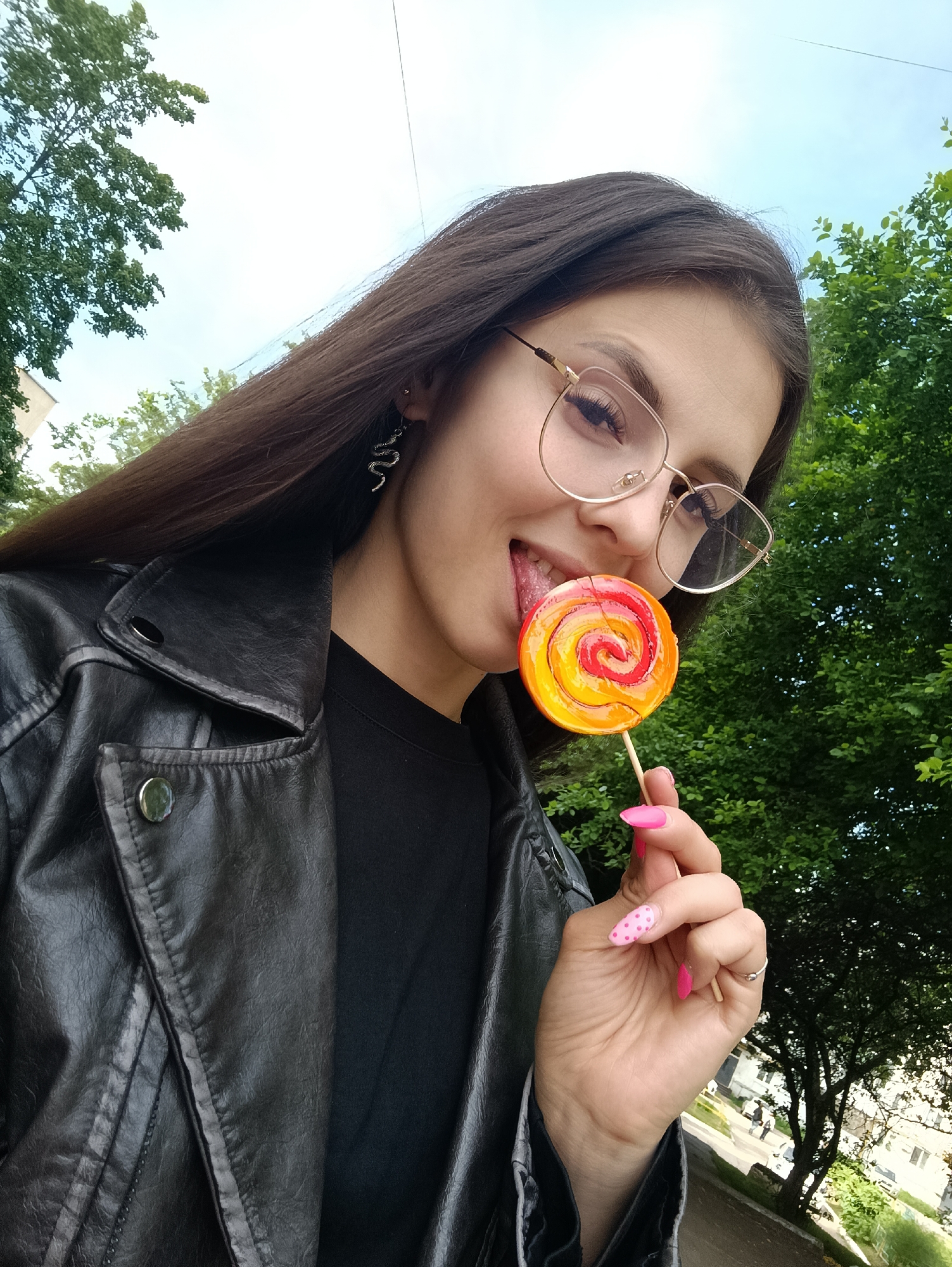 A close-up outdoor selfie features a young woman with long, dark hair and round, gold-rimmed glasses. She is wearing a black faux leather jacket over a black t-shirt. She holds a colorful lollipop with orange, yellow, and red swirls, extending her tongue playfully to lick it. Her fingernails are painted a vibrant pink, with a polka-dot design visible on her right ring finger, which also sports a simple silver ring. A distinctive silver snake-shaped earring hangs from her right ear.
The setting appears to be outdoors during the daytime, indicated by the bright sky and abundant green foliage from trees that form the background. Hints of a building and a paved surface suggest an urban or suburban environment. The overall mood conveyed is playful and casual, with the subject engaging directly with the camera. There is no visible text in the image.