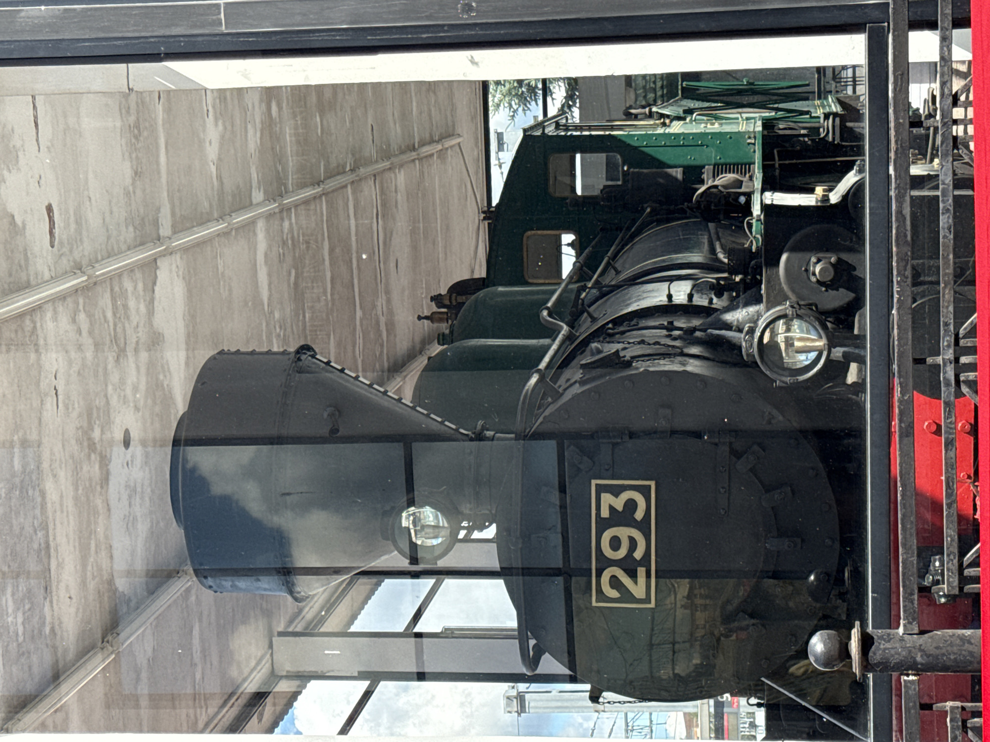 The image shows a steam locomotive, identified by the number "293" on its front, displayed behind a glass enclosure. The locomotive is dark green and black, with a large funnel-like structure at the front. It appears to be stationary and possibly on exhibit. The background shows some architecture, including a concrete ceiling with lights and a glimpse of the sky and trees. Reflections are visible on the glass, obscuring some details. There are no people visible in the image. The locomotive is situated in what appears to be an outdoor or semi-outdoor covered area. There is a railing in the foreground.