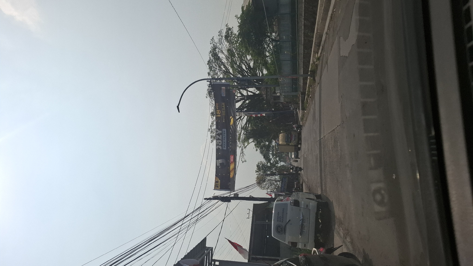 The image captures a street scene in Cikarang, Indonesia, under a bright, hazy sky. In the foreground, the dashboard of a vehicle is visible, reflecting the road and some indistinct structures. The road itself is paved and shows signs of wear, with a slight sheen suggesting recent moisture.
Dominating the mid-ground is a large billboard advertising "JAZY MAX FILTER" with text in Indonesian, including "180,000" and "BIG BESAR". Several utility poles with a dense network of wires run overhead, characteristic of urban Indonesian infrastructure.
On the left side of the road, a white car with the license plate "B 1347 DIX" is parked. On its rear window is a sticker for "BADAN TENAGA NUKLIR NASIONAL" (National Nuclear Energy Agency). A motorcycle is parked behind the white car, and another motorcycle is seen on the road further ahead.
Traffic is moderate, with a truck and several other vehicles moving along the street. To the right of the road, a fenced area and lush greenery, including a large tree, are visible. Indonesian flags, red and white, are also displayed on poles. The overall impression is a typical day on a busy road in a commercial or suburban area of Cikarang, Indonesia.