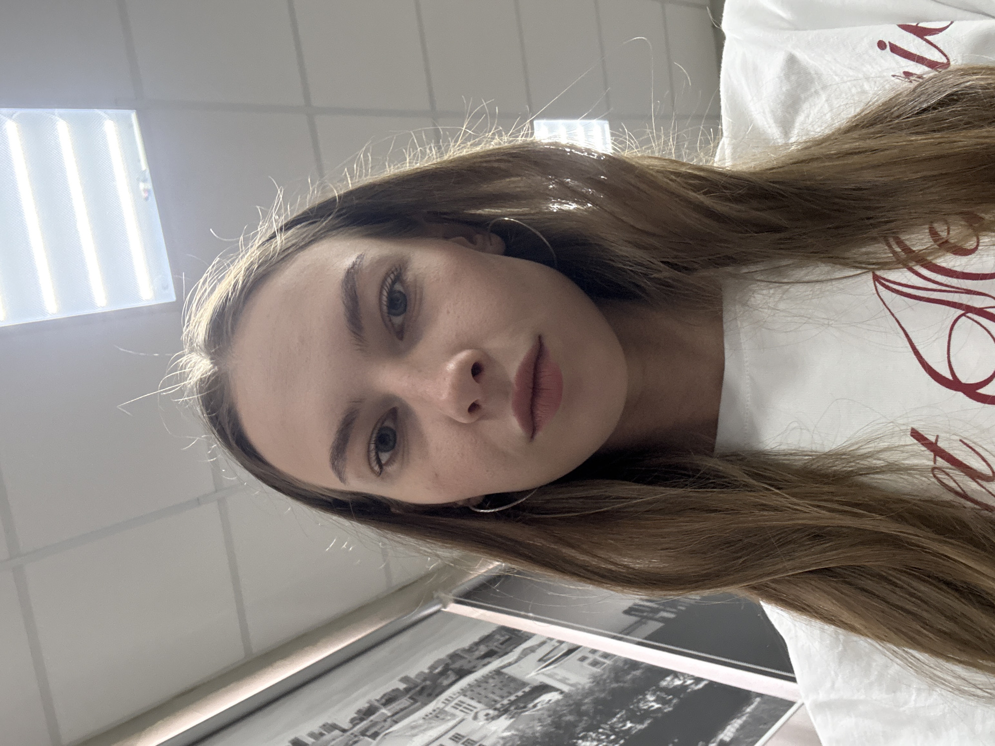 A young woman with long brown hair is looking directly at the camera. She is wearing a white t-shirt with red lettering. The background appears to be an indoor setting with fluorescent lighting on the ceiling and a large black and white mural on the wall to her left. The scene is well-lit. No information is available to determine the location in the city of Perm, Russia.