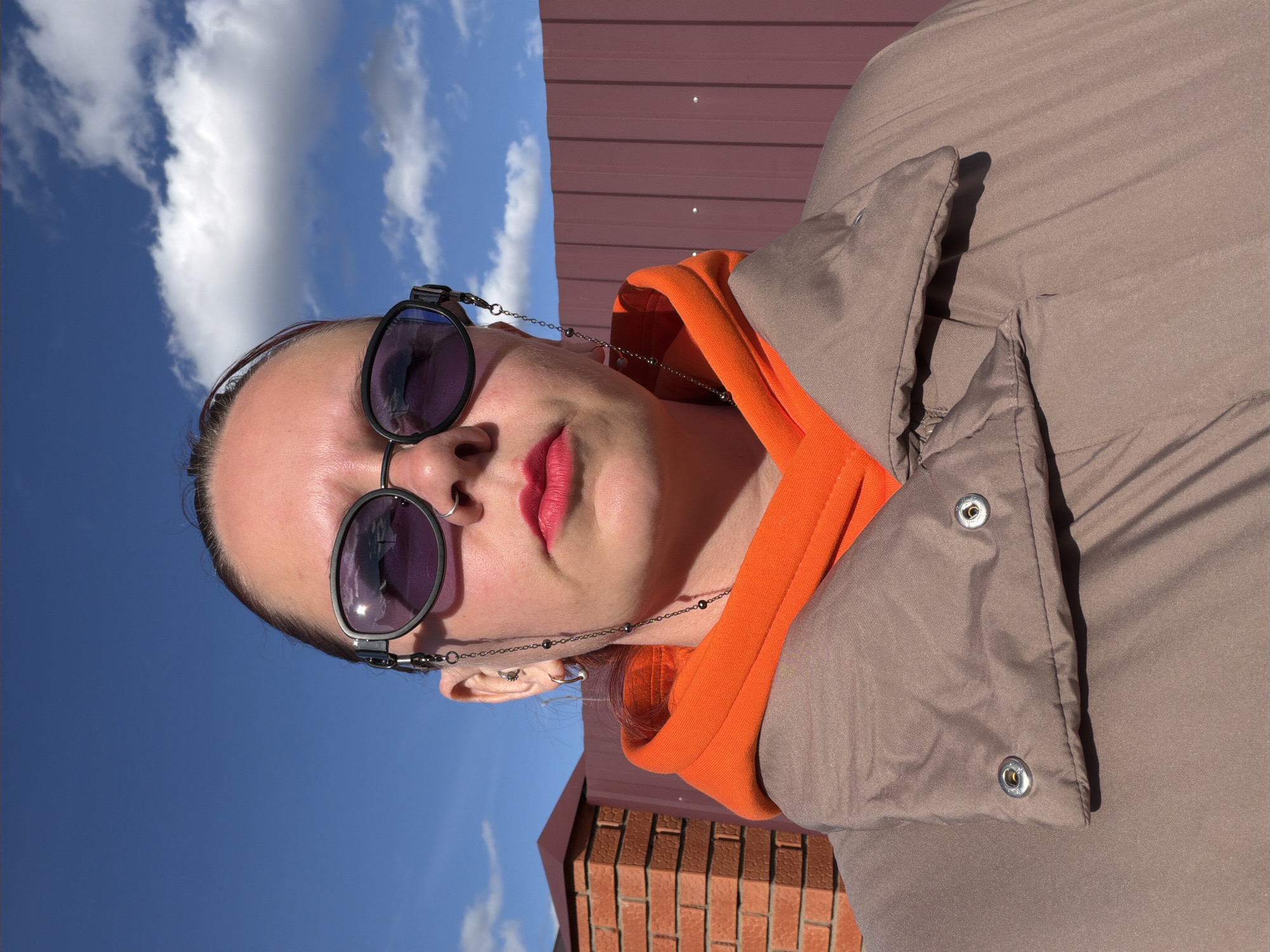 A medium-shot photograph taken outdoors shows a woman with fair skin and dark hair pulled back, wearing sunglasses with a chain, a bright orange hoodie, and a brown puffy jacket. She has multiple ear piercings and is wearing red lipstick. The background includes a clear blue sky with fluffy white clouds, a reddish-brown metal fence, and part of a brick wall. The image is oriented vertically. Based on geolocation metadata, the location is Chaikovsky, Russia.