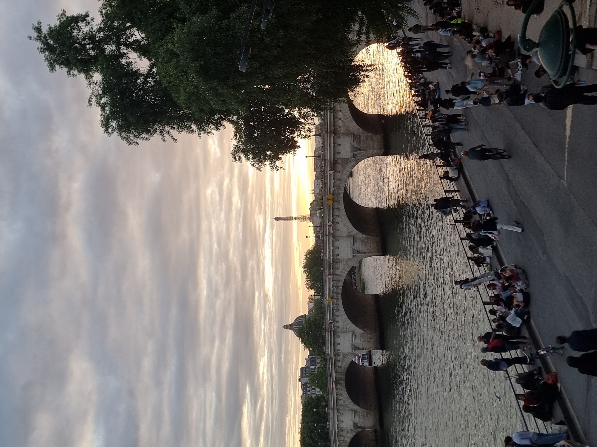 Here's a concise description of the image:
A long bridge with multiple arches spans a river in Paris, France, with the Eiffel Tower visible in the distance under an evening sky. Many people are sitting and standing along the riverbank. The sky features striations of gray and pale light. The water reflects the light. A dark tree branch is visible in the upper left. A streetlight is in the upper right.