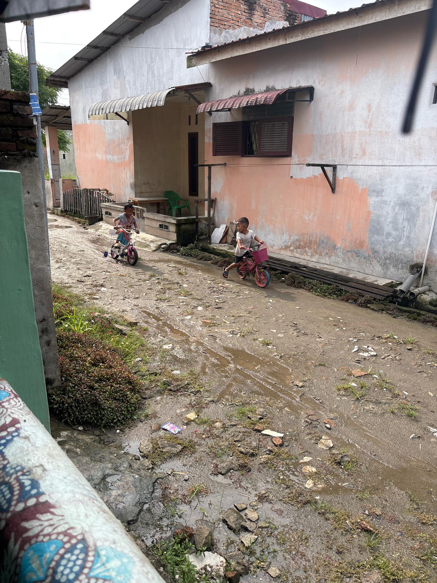 Two children are riding bicycles down a muddy, unpaved alleyway between two weathered buildings. The building on the right has a salmon-pink lower half and a gray upper half, with a corrugated metal awning over a window. The building on the left has a similar salmon-pink lower half and a lighter gray upper half, with a metal awning over what appears to be a doorway. In the foreground, a green pillar with a patterned fabric is visible. The ground is uneven, with puddles of water, debris, and sparse vegetation. The sky is overcast. The scene is likely in a residential area of Helvetia, Indonesia, characterized by its simple architecture and unpaved roads.