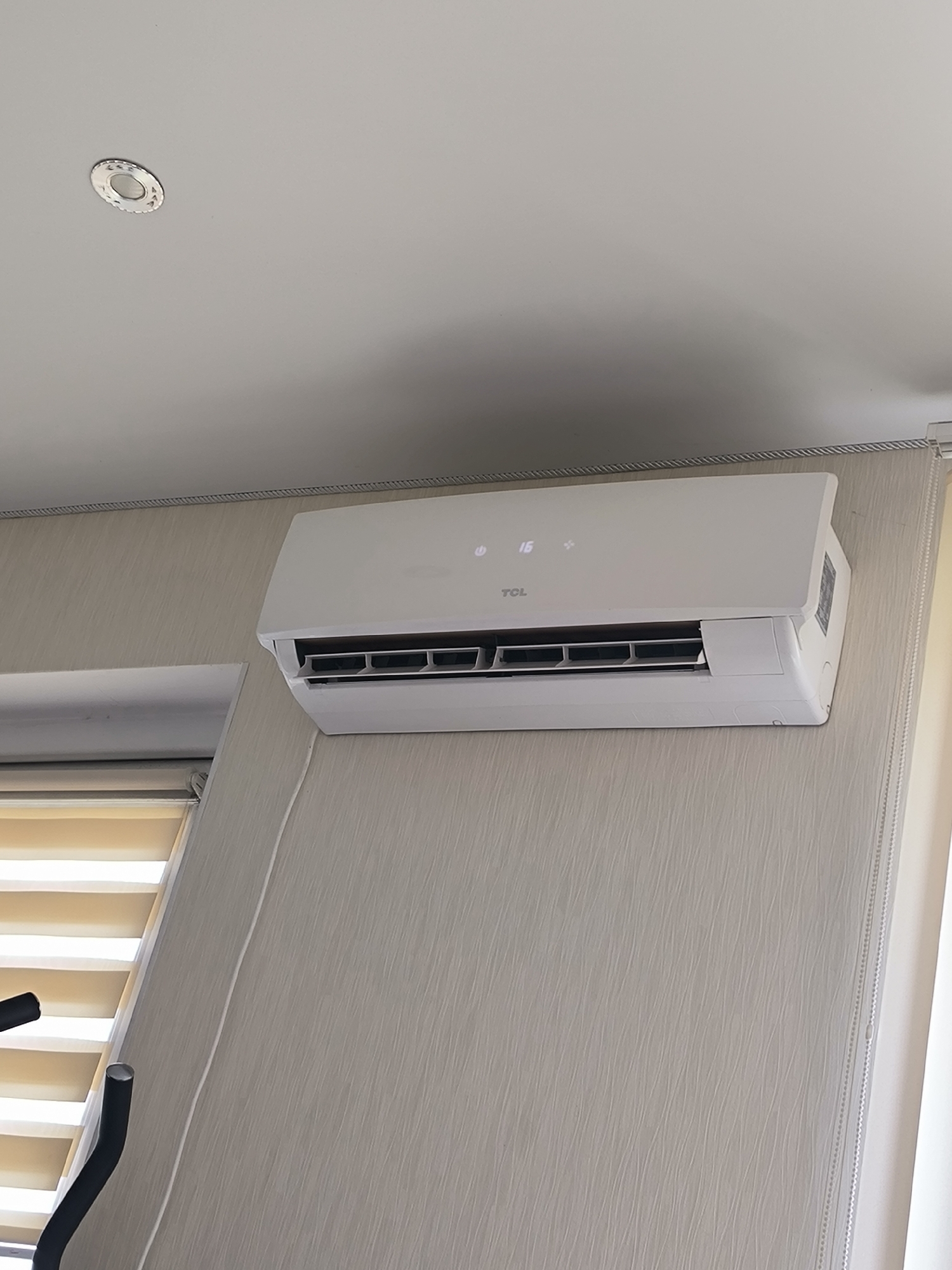 A white TCL brand air conditioner is mounted high on a wall with textured, light beige wallpaper. The air conditioner has a digital display showing "16" degrees and symbols for power and fan speed. Below the air conditioner, a set of horizontal blinds with alternating opaque and translucent panels are partially visible. To the left of the blinds, two black metal support arms from exercise equipment are in the lower left corner of the frame. The ceiling above is white with a circular recessed light fixture. The image is taken from a medium-low angle, looking slightly upwards. This scene is likely from an indoor residential setting in Chișinău, Moldova, given the visual cues and common interior design elements.