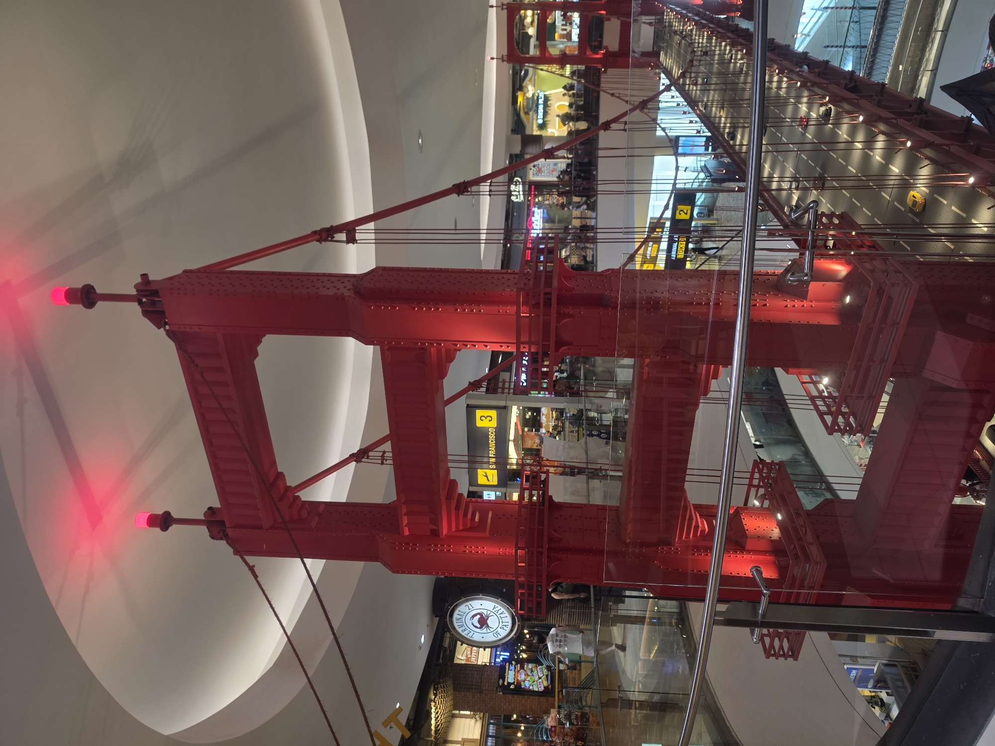 Here is a forensic description of the image:
The photograph appears to be taken from a lower vantage point, looking upwards at a large-scale replica of the Golden Gate Bridge, painted in red. The replica spans multiple levels inside what seems to be a shopping mall or indoor commercial space.
The bridge’s structure is visible in detail, including the supporting towers, cables, and road surface, which has miniature cars on it. Red lights are affixed to the top of the towers. The background reveals the interior architecture of the mall, with multiple levels of shops and walkways. Some of the shops include signs and branding. The location is identified via signage as Pattaya City, Thailand, specifically Terminal 21 Pattaya. A large clock is seen below the bridge's structure with the lettering "TERMINAL 21 OF PATTAYA". Also present are signs to SAN FRANCISCO. People are indistinctly visible in the background shopping and walking.