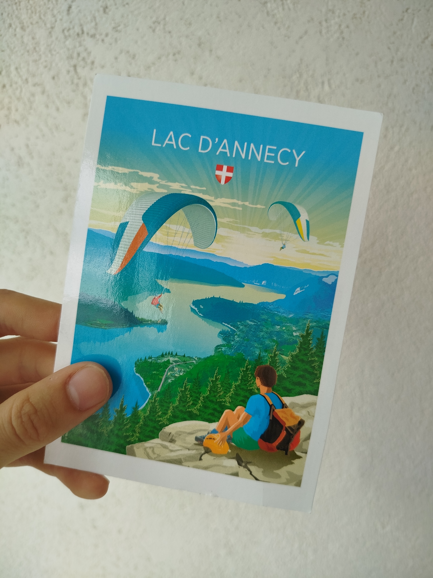 A person is holding a postcard featuring an illustration of Lac d'Annecy, France. The illustration shows two paragliders flying over the lake, mountains, and trees, with a person sitting on a rock in the foreground. The text "LAC D'ANNECY" is printed at the top, along with a Swiss flag. The background is a white wall with a textured surface.