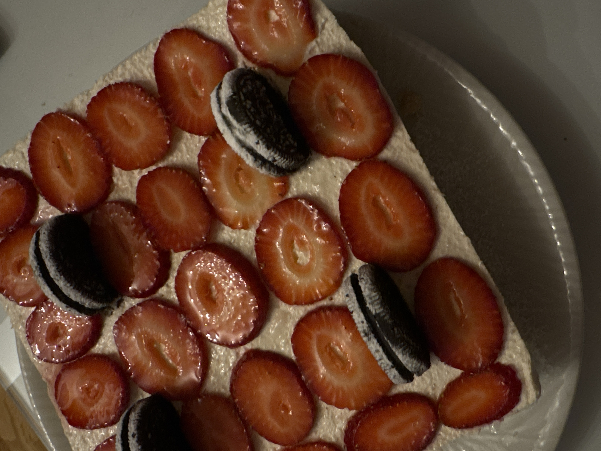 A close-up shot depicts a square-shaped dessert, possibly a cake or tart, adorned with sliced strawberries and Oreo cookies. The dessert is placed in a round, shallow dish with a white or light-colored interior and a darker rim. The strawberries are arranged closely together, covering the majority of the dessert's surface. The Oreo cookies are interspersed among the strawberries, seemingly pressed into the dessert's cream layer. The dessert itself has a light-colored creamy or custard-like layer visible between the strawberries. The scene's ambient lighting appears somewhat dim and soft. The location in the city of Bar, Montenegro is unknown.