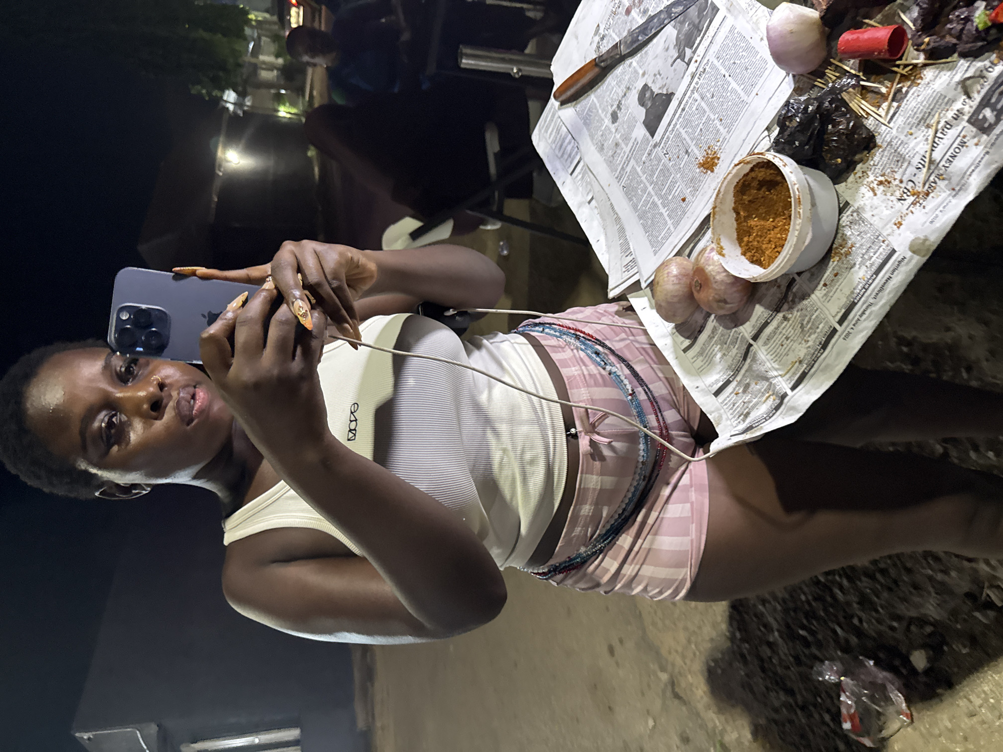 A Black woman is holding up a smartphone to take a picture of herself outdoors at night in Abuja, Nigeria. She is wearing a white ribbed tank top, pink and white striped shorts, and a beaded accessory around her waist. She has long, decorated fingernails. In front of her, on a spread of newspapers, are two onions, a small container of brown spice, and skewers of meat. A knife is also on the newspapers. Other people and structures are visible in the blurred background.