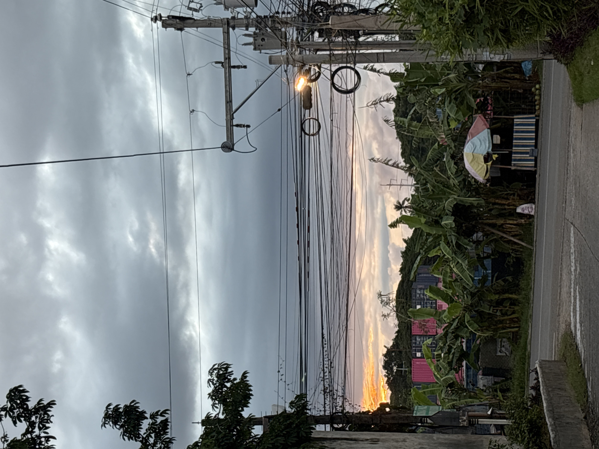 The image captures a street scene in Davao City, Philippines, at dusk. The sky is overcast with dramatic clouds, hinting at an approaching storm, but a sliver of orange sunset peeks through the horizon, illuminating the clouds from below. In the foreground, a road runs parallel to a concrete barrier and a patch of greenery. To the left, a weathered concrete structure with some hanging items is visible, along with the dark silhouette of a tree.
Dominating the midground is a dense cluster of banana trees, their large leaves creating a lush, tropical backdrop. Behind the banana trees, several shipping containers are stacked, painted in vibrant pink and blue. A small roadside stall with a multi-colored umbrella and striped awning is set up amidst the banana trees, with several green coconuts displayed in front.
The right side of the image is dominated by a utility pole cluttered with numerous electrical wires and transformers. A street lamp on the pole is illuminated, casting a warm glow. The overall impression is one of a bustling, yet somewhat chaotic, urban fringe in Davao City, characterized by a mix of natural elements, industrial shipping containers, and everyday street life, all under a brooding sky.