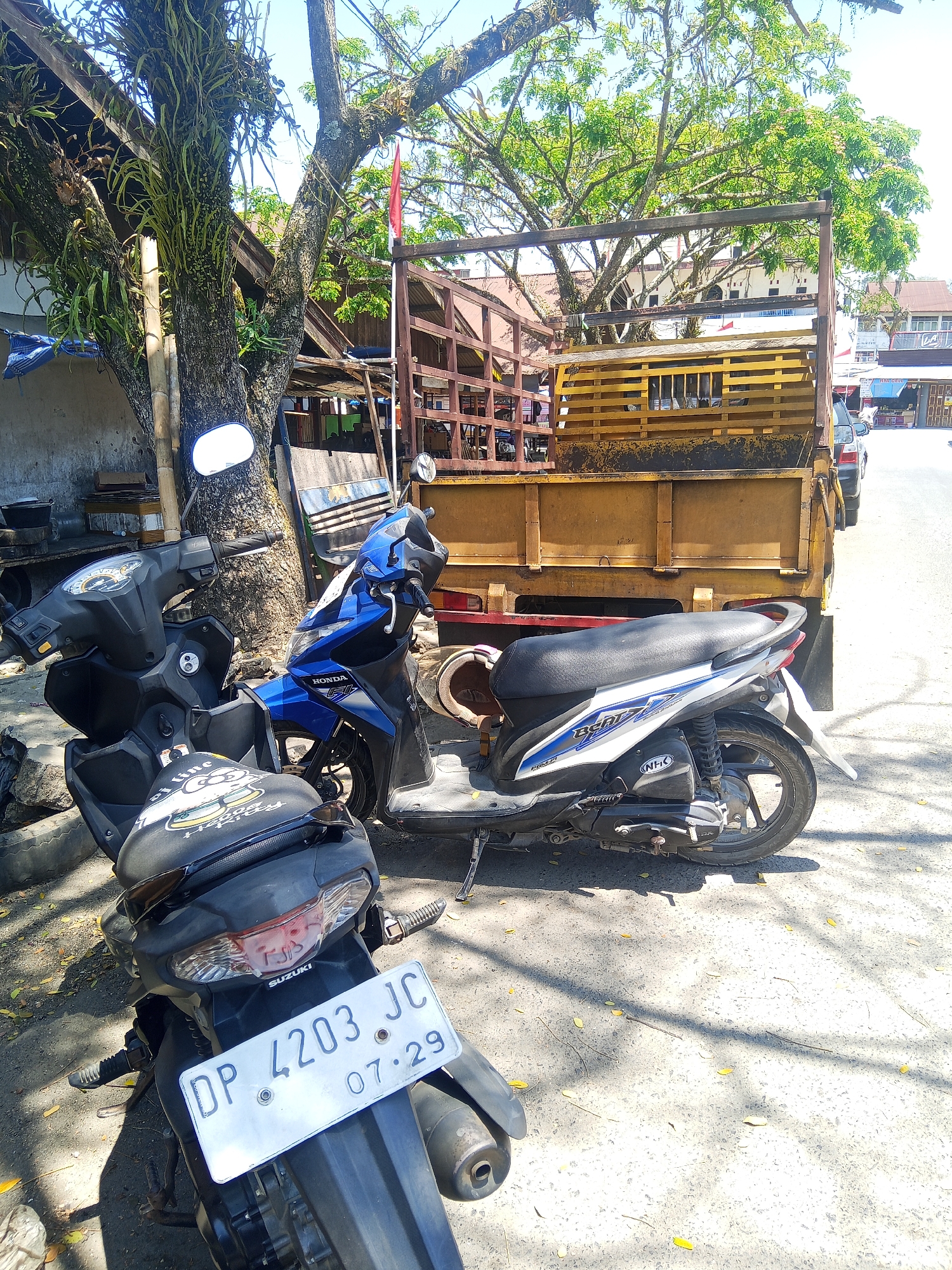 Two scooters are parked on a paved surface, with a large truck in the background. The scooter in the foreground is black with a decorative seat cover featuring a cartoon character. Its license plate reads "DP 4203 JC 07.29". A blue and black Honda scooter is parked to the right of the black scooter. Behind the scooters is a wooden truck with a metal frame. A large tree with abundant foliage is to the left of the truck. The scene appears to be in Makale, Indonesia, as suggested by the style of the license plate and the general environment. The sky is clear and blue, indicating a sunny day. There are no people visible in the image.