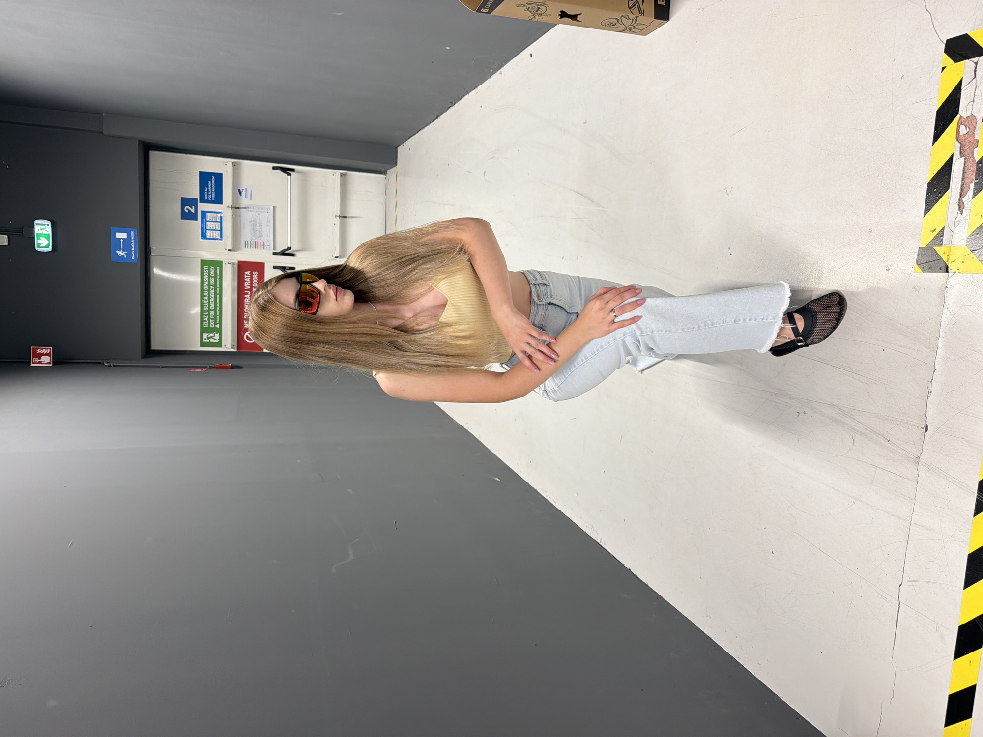 A young woman with long blonde hair and red-tinted sunglasses kneels on a white floor in a narrow hallway with gray walls. She wears a tan crop top and light blue jeans with frayed hems. Her hands are clasped in front of her. Behind her, a double door is marked with various signs, including an exit sign and a sign with the number "2." The hallway is illuminated by overhead lights, casting shadows on the floor. Yellow and black hazard tape is visible on the floor in the foreground and in the background. A cardboard box with markings is positioned to the right of the woman.