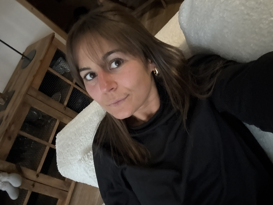 A woman with shoulder-length brown hair and bangs poses for a selfie. She wears a black turtleneck and gold hoop earrings. Her expression is neutral. To her left is a wooden cabinet with wire mesh doors. A fluffy white object, possibly a cushion or part of furniture, sits behind her head. The overall lighting is soft. The photo appears to be taken indoors.