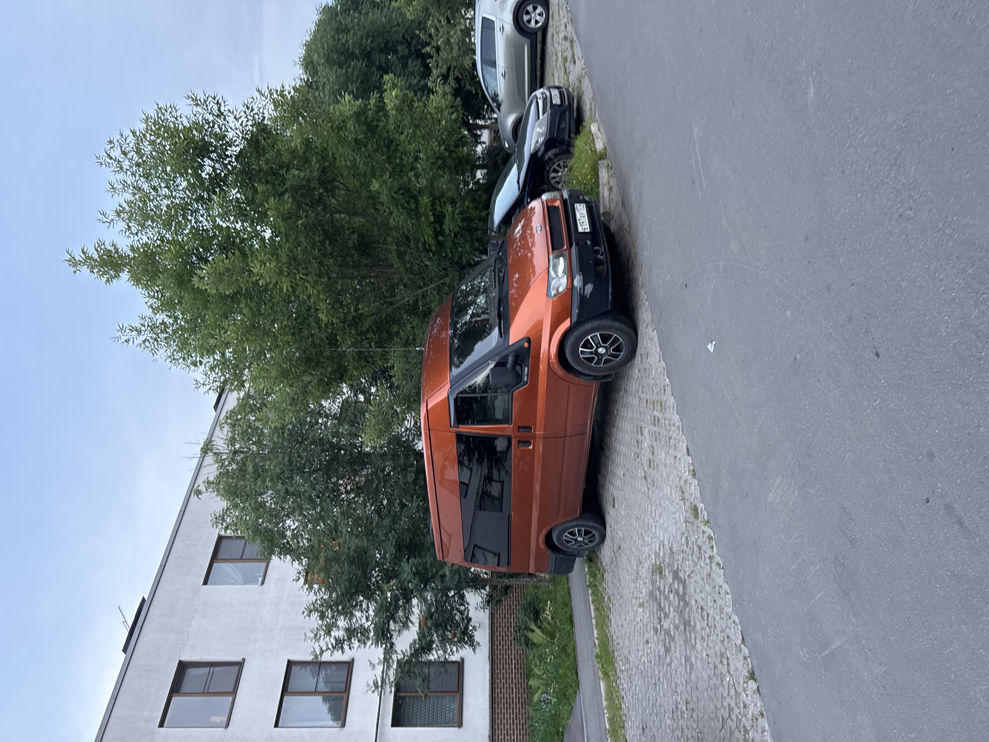 An orange van is parked on a paved area next to a modern white apartment building in Domodedovo, Russia. The van is a Ford Transit and has tinted windows. To the right of the van, a black car and a silver SUV are also parked. Trees and green foliage are visible behind the parked vehicles. The sky is overcast, suggesting it is either early morning or late evening. The area appears to be a residential parking lot.