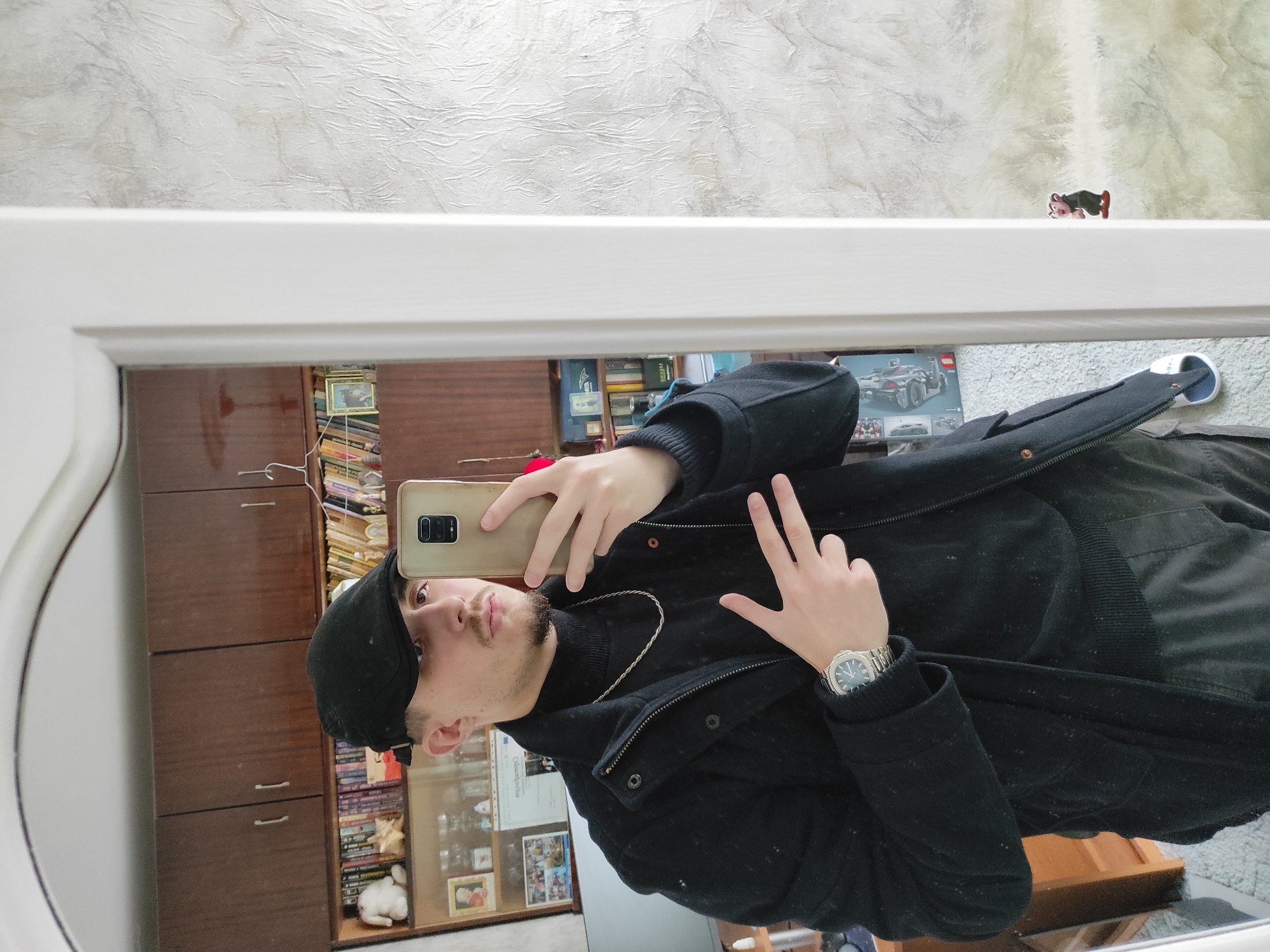 A man is taking a selfie in a mirror. He is wearing a black beanie, a black turtleneck, and a black jacket. He is holding a phone in his left hand and making a peace sign with his right hand. He is also wearing a watch on his left wrist. The reflection in the mirror shows a room with a shelf filled with books and other objects. There's also a small figurine of a cartoon character on the top of the mirror frame. The location is not discernible as Varna, Bulgaria, based solely on the visual information provided in the image.