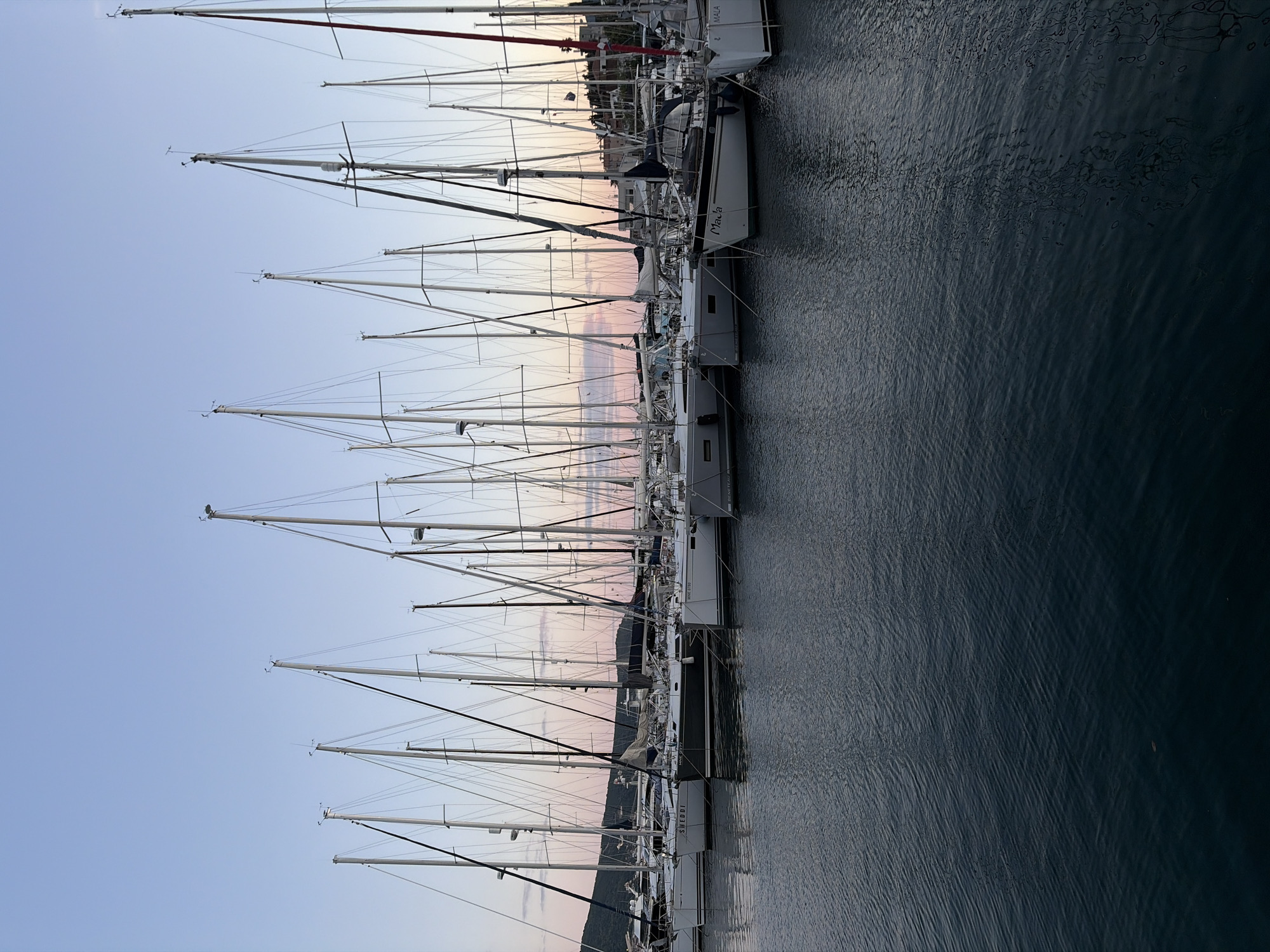 This image, captured in Meljine, Montenegro, presents a serene marina scene, likely at sunset or sunrise. The photograph is oriented vertically, showing numerous sailboats neatly docked side by side along a quay.
The foreground is dominated by the dark, calm water of the marina, with subtle ripples and reflections visible on its surface. Moving across the image, the hulls of the sailboats are visible, predominantly white or light-colored, with dark accents. Several boat names and manufacturers are legible on the hulls, including "MALA," "SHEDDI," "BENETEAU," and "PINE 9180."
Above the hulls, a dense array of tall, slender masts and intricate rigging lines stretches upwards. Many masts feature wind indicators and other navigational equipment at their tips, silhouetted against the sky.
The sky, occupying the upper portion of the image, displays a beautiful gradient of colors. It transitions from a light, clear blue at its highest point to warm, soft hues of pink and orange closer to the horizon, indicative of the golden hour. A faint outline of land, possibly a hillside with some distant buildings or trees, can be seen along the horizon line, nestled between the sky and the boats.
The overall scene conveys a sense of tranquility and peace. There are no people clearly visible or any distinct activities taking place, emphasizing the quiet stillness of the harbor at this gentle hour.