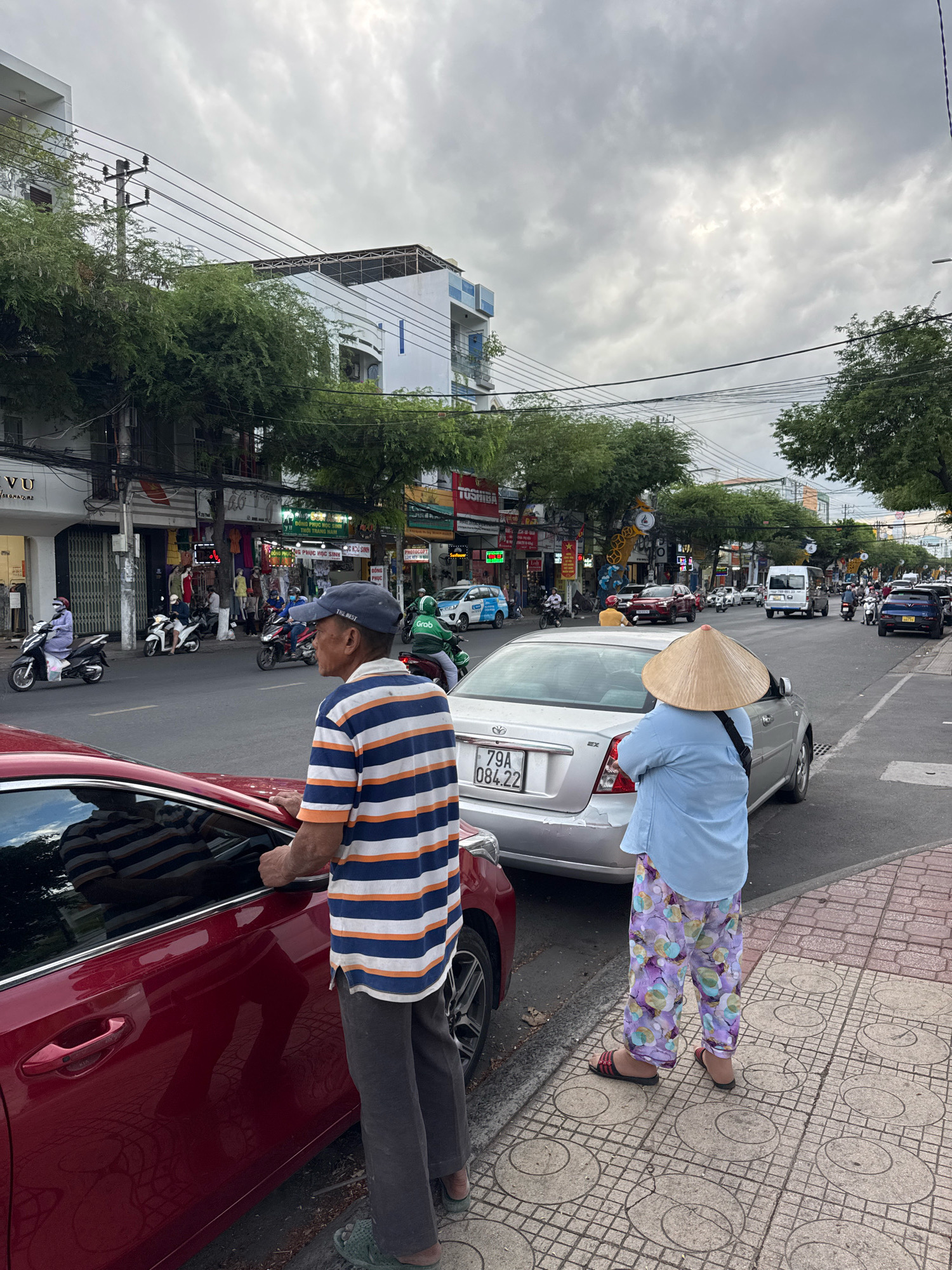 The image depicts a street scene in Nha Trang, Vietnam, on a cloudy day. In the foreground, a man wearing a blue and orange striped t-shirt and dark pants stands next to a red car. To his right, a person wearing a light blue shirt and colorful patterned pants, with a conical hat on their head, stands on the sidewalk. A silver sedan is parked behind the red car. The street is lined with buildings and trees, and traffic, including motorcycles and cars, is visible in the background. The sidewalks are paved with tiles, and overhead power lines crisscross the sky. The scene appears to be a typical urban environment in Vietnam, with active street life and commerce.