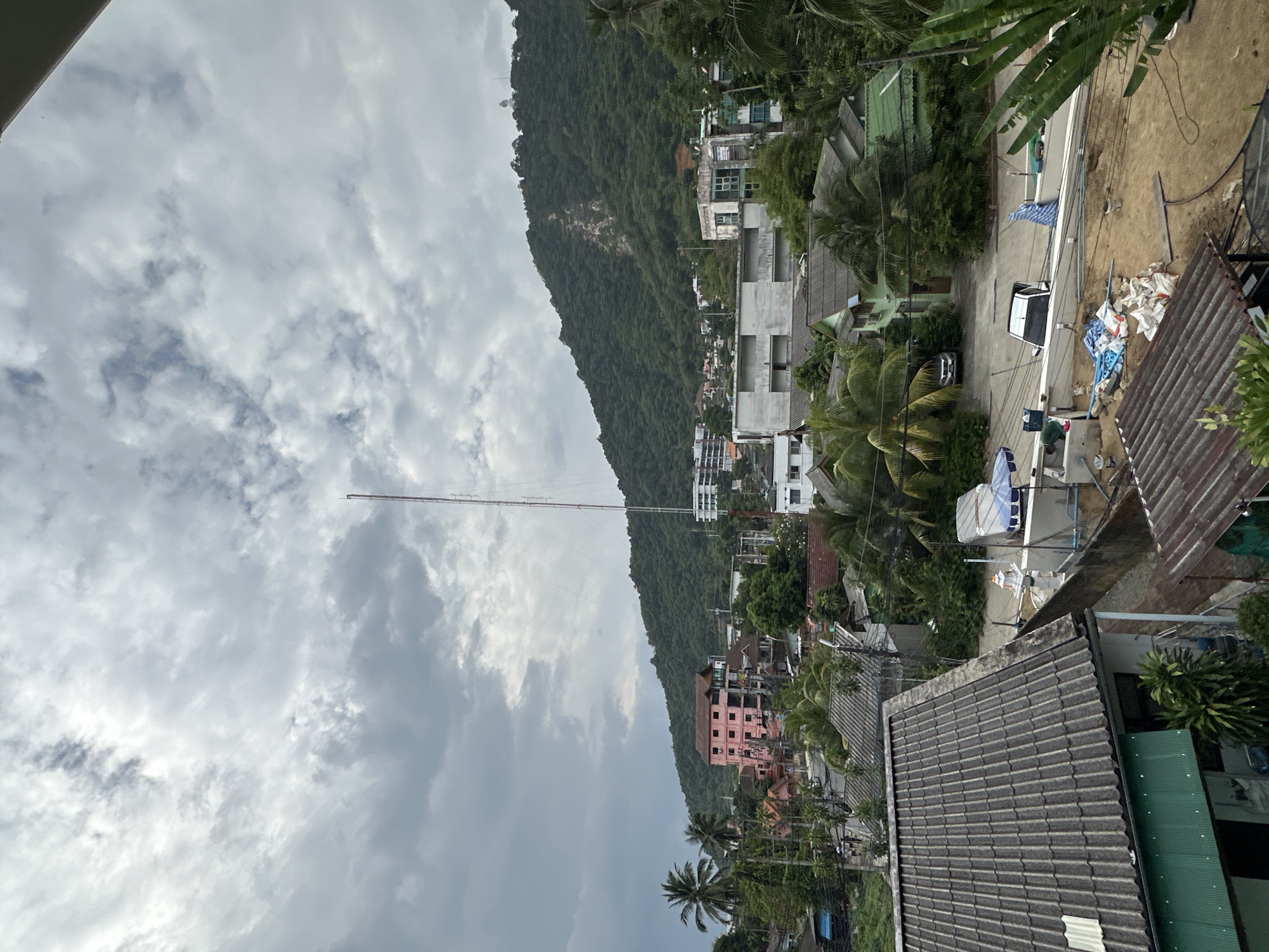This image captures a panoramic view of Karon, Thailand, characterized by a mix of residential buildings, lush greenery, and a prominent mountainous backdrop. The sky is overcast with dense clouds, suggesting a day with potential for rain. In the foreground, a grey tiled roof of a residential building is visible, with a green corrugated metal roof and a lush green plant in the lower left corner.
Moving further into the scene, we see a variety of buildings, including a striking pink multi-story structure that stands out against the muted tones of its surroundings. Interspersed among the buildings are numerous palm trees, a characteristic feature of tropical landscapes. A tall communications tower pierces the sky in the mid-ground, signifying infrastructure within the town.
The background is dominated by a large, verdant mountain, its slopes covered in dense foliage. A light-colored structure, possibly a monument or building, is visible near the peak of the mountain. The overall impression is that of a developing urban area nestled within a natural, tropical environment. The ground level of the image shows a construction or renovation site, with piles of sand, building materials, and tarps suggesting ongoing work. Two cars are parked in the area, one white and one black, and a blue and white striped umbrella is set up near some construction materials.