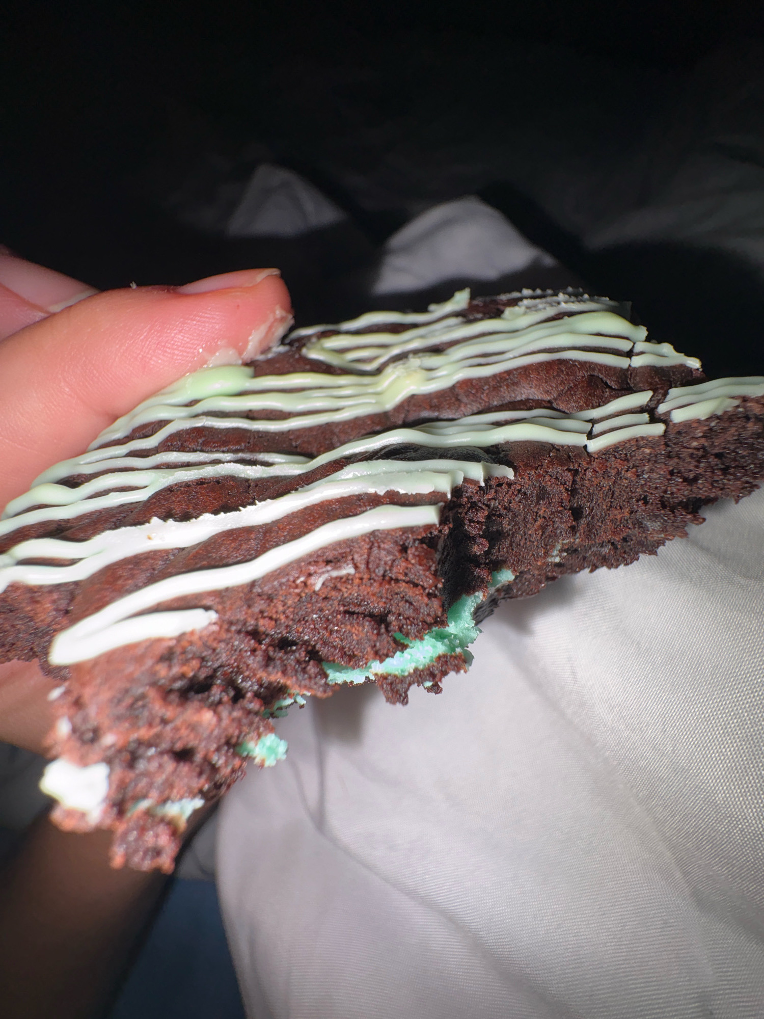 A close-up, angled shot shows a hand holding a piece of chocolate brownie with a mint-green filling and white and mint-green drizzled icing. The brownie is dark brown and appears moist and dense, with visible crumbs and a rough texture. The icing is applied in thin, wavy lines. A bite has been taken out of the brownie, revealing the bright mint-green filling underneath. The hand holding the brownie has a light skin tone and appears to be a thumb and forefinger. The background is dark and out of focus, with some lighter, indistinct shapes that could be bedding or fabric. The scene is not set in a city.