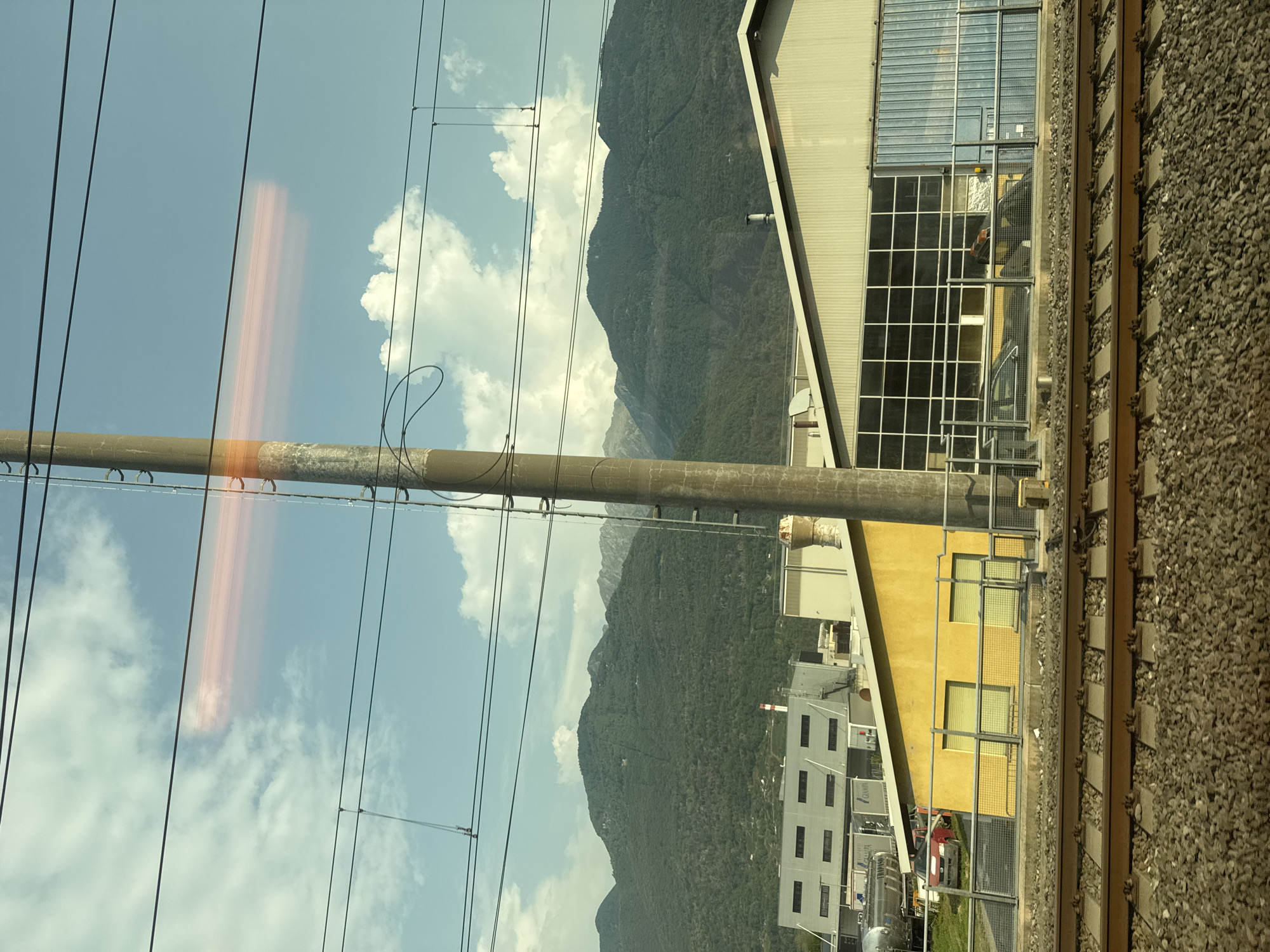 The image, taken from a moving train, shows a view of industrial buildings and a mountainside. In the foreground, railroad tracks run horizontally across the bottom of the frame. Beyond the tracks, there is a yellow building with two windows and a large industrial building with a light-colored facade and a section with large windows. In the background, a densely forested mountain range rises against a partly cloudy sky. A tall, slender pole with overhead wires runs vertically through the center of the image. A horizontal orange light streak is visible in the sky, likely a lens flare. The scene appears to be in an industrial area at the base of a mountain, with clear skies and daylight.