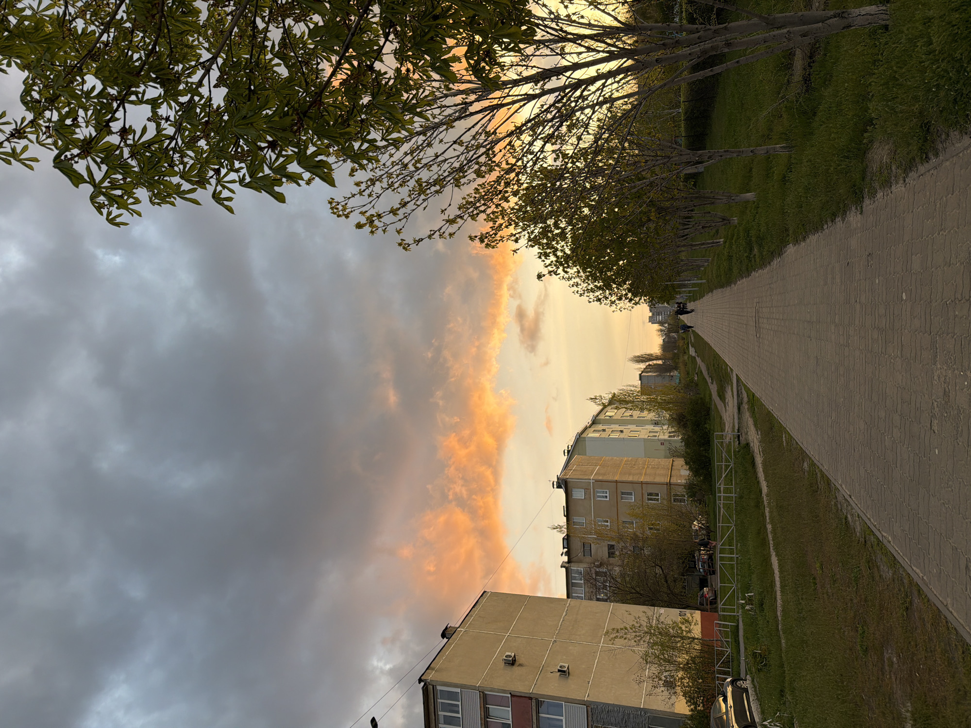 The image captures a pedestrian walkway in what appears to be a residential area in Kamyshin, Russia. The scene is framed by trees with emerging foliage both on the left and right.
The walkway is paved with light-colored bricks and extends into the distance, where a few individuals are visible. Adjacent to the walkway is a strip of green grass.
To the left of the walkway, several multi-story buildings are present. The buildings are constructed of concrete and have rows of windows, some with air conditioning units. A car is parked near the buildings.
The sky is filled with dramatic clouds, with a mixture of dark gray clouds to the left and vibrant orange hues near the horizon, likely due to the setting sun. The lighting gives the scene a warm, golden ambiance.
There are no visible interactions or activities, but the scene conveys a sense of urban tranquility during twilight.