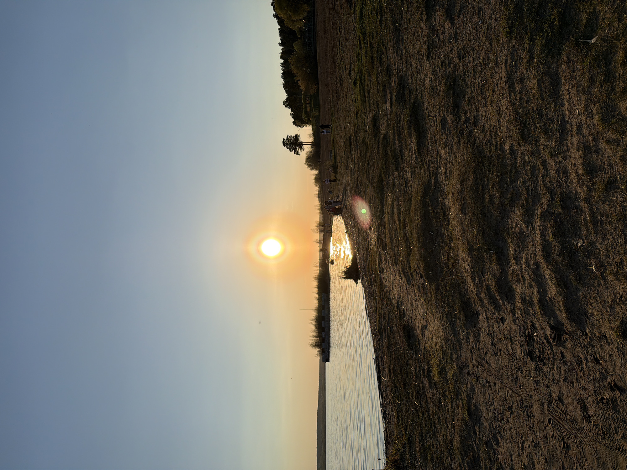 The image shows a landscape during sunset. The sun is setting on the horizon, casting a bright light and reflecting on the water's surface. The sky is a gradient of blue to orange. A shoreline is visible, with a sandy area on the right. The land appears to have grass and sparse vegetation. In the distance, there are silhouettes of trees. Some indistinct figures of people are visible. The location might be a bank of the Izhevsk pond, possibly near the city of Izhevsk, Russia.