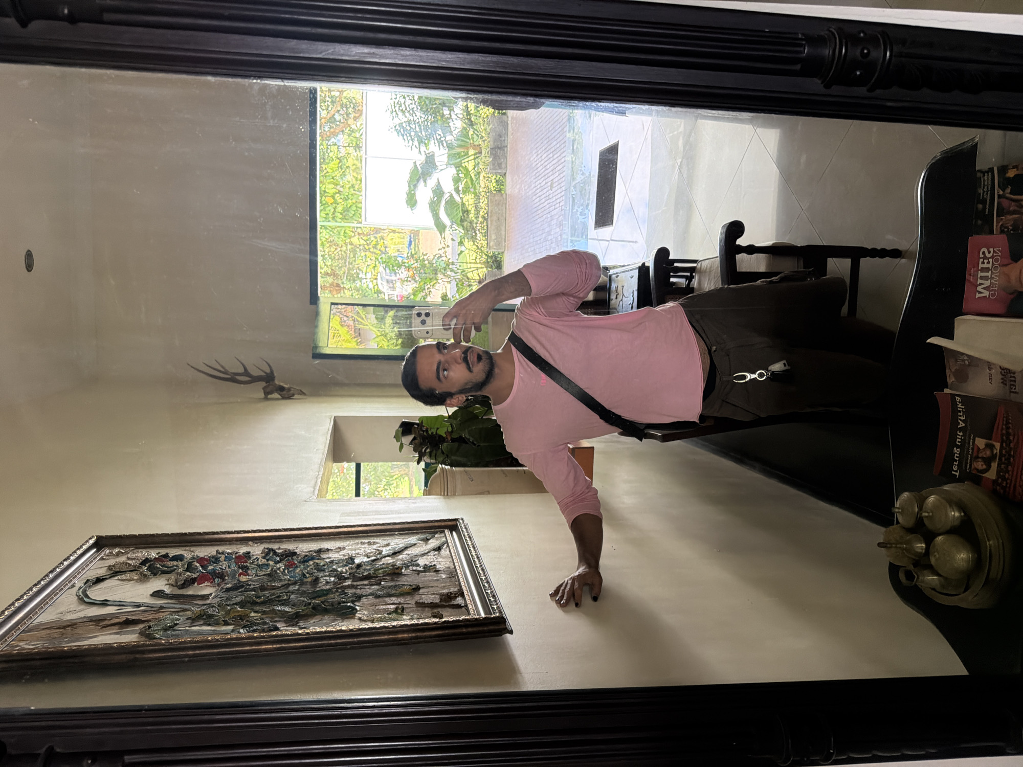A man with a beard and dark hair, wearing a pink long-sleeved shirt and brown pants, stands in front of a large mirror, taking a selfie. He has a black strap across his chest. His left hand is resting on a cream-colored wall, adorned with a framed artwork. To his right, the mirror reflects an open doorway leading to an outdoor area with lush greenery and trees. In the foreground of the mirror's reflection, there are two dark wooden chairs. The overall scene appears to be an indoor space with a view of a garden or courtyard.