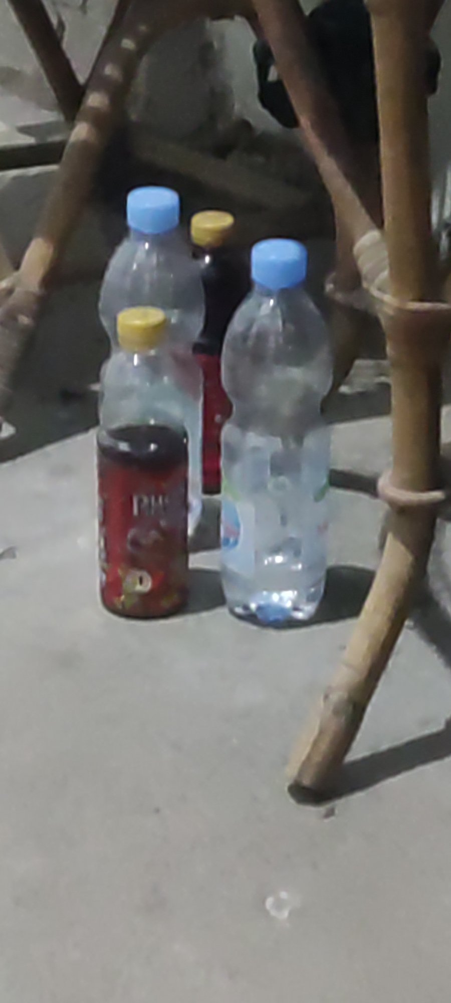 The image shows several plastic bottles and a can of soda on a concrete surface. The bottles are of various sizes and colors, with some filled with clear liquid and others with darker liquid. One bottle has a blue cap, another a yellow cap, and a third also has a blue cap. A red can with white lettering and a cartoon image of a dog is visible in the foreground. The background is out of focus but appears to be a blurry interior or exterior setting with wooden structures. The lighting suggests it is either evening or an indoor dimly lit area. The scene is static and no people are visible. The location in Karangwangun, Indonesia, is not discernible from the visual information presented in the image.