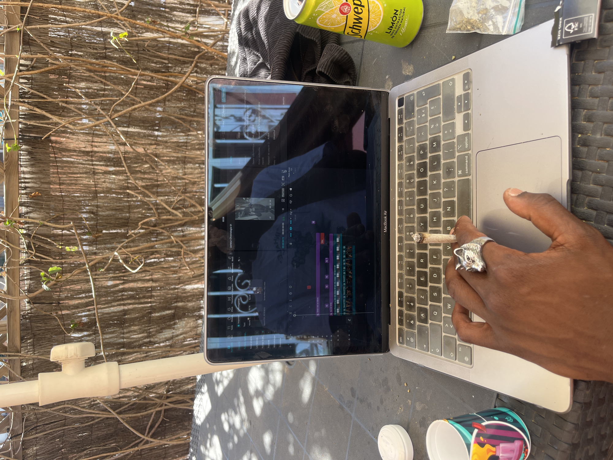 A person is working on a MacBook Air laptop outside, holding a lit cigarette between their fingers while typing. The laptop displays video editing software with timelines and visual elements. Beside the laptop is a can of Schweppes Lemon soda, a small plastic bag containing green material, a packet of rolling papers, and a colorful cup. The person is wearing a silver ring in the shape of an animal head. In the background, there is a wicker-like fence and a white PVC pipe. The location is likely outdoors, as suggested by the natural lighting and the fence. It's difficult to ascertain the exact location in Barcelona, Spain without additional context.
