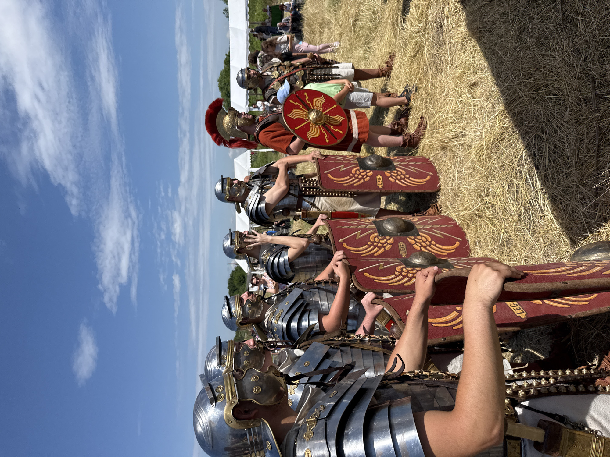 The image depicts an outdoor historical re-enactment event in a dry, grassy field under a clear blue sky with scattered white clouds. The location is Gigen, Bulgaria.
Seven adult males dressed in Roman military attire are arranged in a line, extending from the left foreground into the mid-ground, generally facing towards the right of the frame. From left to right:
The first three re-enactors wear silver-toned lorica segmentata armor and Imperial Gallic helmets with brass accents. They hold tall, curved, rectangular scuta shields, dark red with yellow stylized wings or lightning bolt motifs and the letters "LEG" inscribed. The leftmost re-enactor's bare right forearm is visible, resting on his shield.
The fourth re-enactor, identifiable as a centurion, wears a reddish-brown tunic, a gold-colored helmet adorned with a prominent red transverse crest, and holds a circular red shield (parma or clipeus) featuring a central gold boss and radiating spikes or laurel leaves. His left hand is placed on the shoulder of a young boy standing to his right. The boy wears a light blue baseball cap, a light green t-shirt, and shorts.
The fifth and sixth re-enactors wear silver-toned lorica segmentata armor and helmets similar to the first three, positioned behind the boy and the centurion, respectively.
The seventh re-enactor, on the far right, also wears similar armor and helmet, with his left hand raised towards his face.
The ground consists of dry, yellowish-brown hay or straw, indicating a harvested field or parched grass. A sharp, dark shadow diagonally bisects the lower right foreground. In the background, several white canopy tents are visible, suggesting an event or festival. Numerous other individuals, some observing and others seated, are present near the tents. Green foliage, likely trees or bushes, is visible in the distance behind the tents. The scene is brightly lit by natural sunlight.