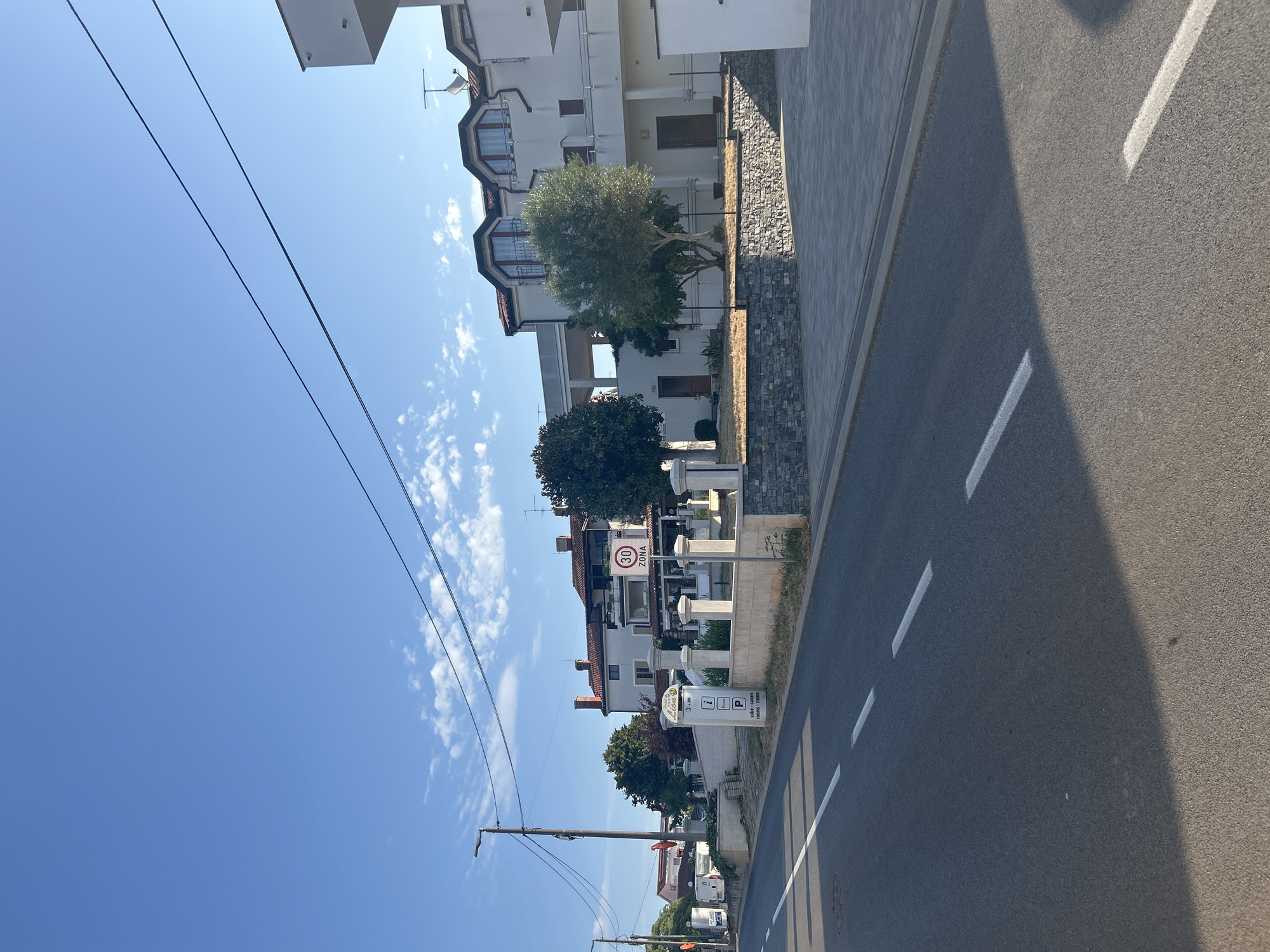 This image captures a street scene in Zambratija, Croatia. The foreground shows a paved road with white dashed lines indicating lanes. A shadow from an unseen object stretches across the road. To the right, a neatly paved area leads to a low stone wall and a sloped embankment. Beyond the embankment, a modern white building with multiple levels and balconies is visible, featuring several windows and roof antennas. Adjacent to this building is another white structure with a distinct balcony. In the middle ground, a signpost with information symbols, including an "i" for information and a "P" for parking, stands near a rounded pillar. A circular speed limit sign indicating "30 ZONA" is also present. Several trees, including an olive tree, are scattered throughout the property. The background is dominated by a clear blue sky with a few wispy clouds and overhead power lines. A street lamp is visible on the left side of the image. The scene appears to be a residential or commercial area in a sunny climate.