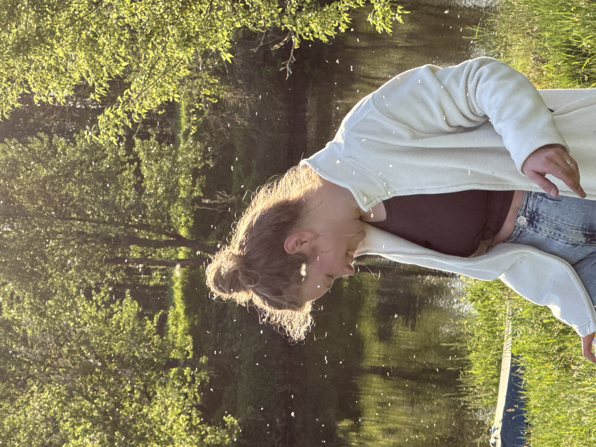 A young woman stands outdoors near a body of water. She is wearing a brown crop top, blue jeans, and a light-colored cardigan. Her hair is styled in a bun. There appear to be small, light-colored particles suspended in the air, possibly insects or pollen. The background includes trees and water, suggesting a park or natural area. The photograph is taken in Egorii, Russia.