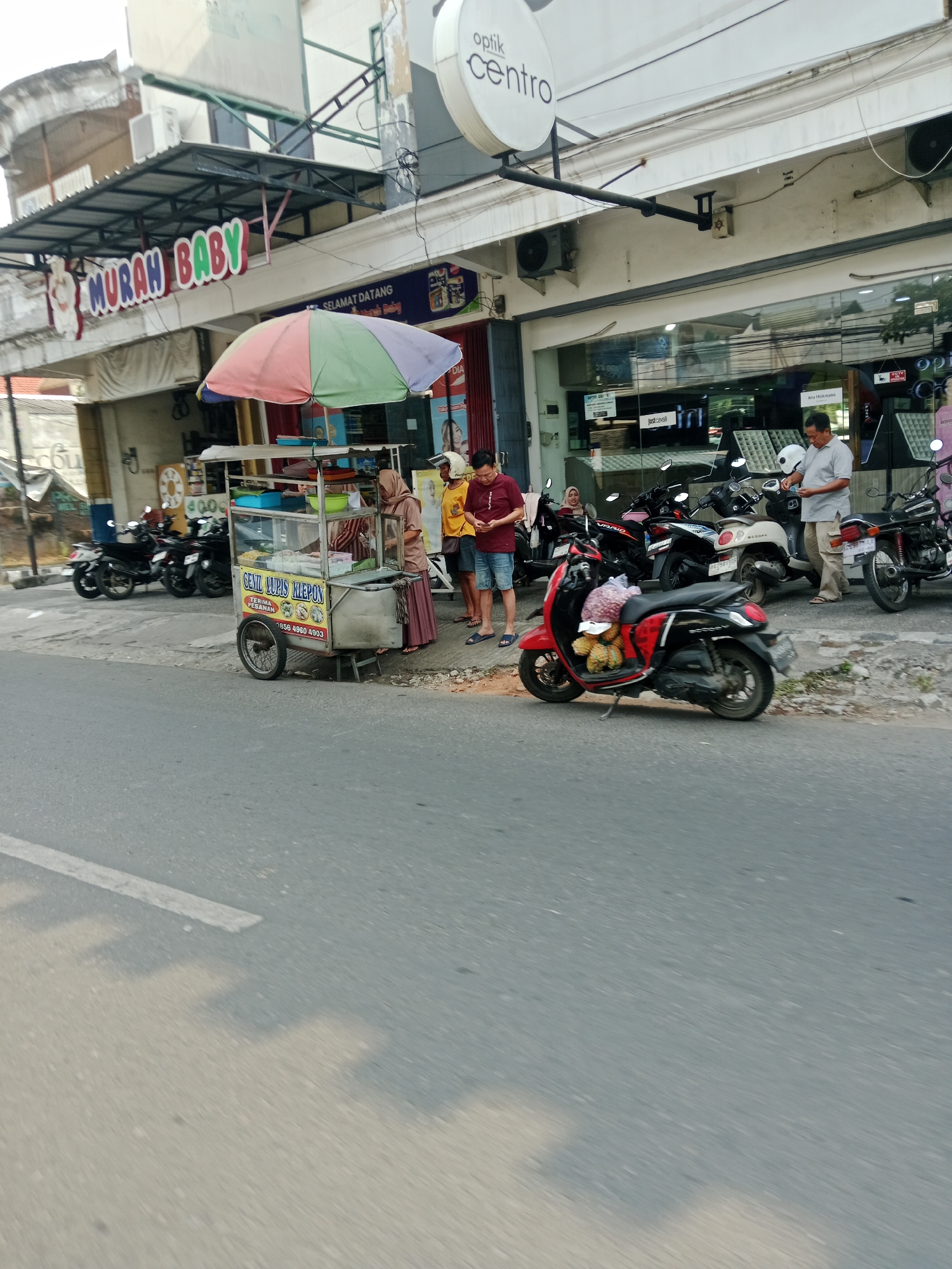 The image shows a street scene in Kediri City, Indonesia. In the foreground, a paved road with a white line is visible. On the right side of the road, a sidewalk is lined with parked motorcycles. A red and black scooter is parked in the foreground with a bag of fruit on its seat. Behind the scooter, a food cart is set up under a multi-colored umbrella. The cart has signage that reads "GENIL JURUS KIEPON" and "TERIMA PESANAN". Three people are standing near the food cart: a woman wearing a hijab and a long skirt, a man in a yellow shirt and shorts, and another man in a red shirt and shorts. To the left of the food cart, there are more parked motorcycles. In the background, a building with a sign that says "MURAH BABY" is visible. Further to the right, a building with a sign that says "optik centro" is present. The overall scene depicts a commercial street with food vendors and shops.