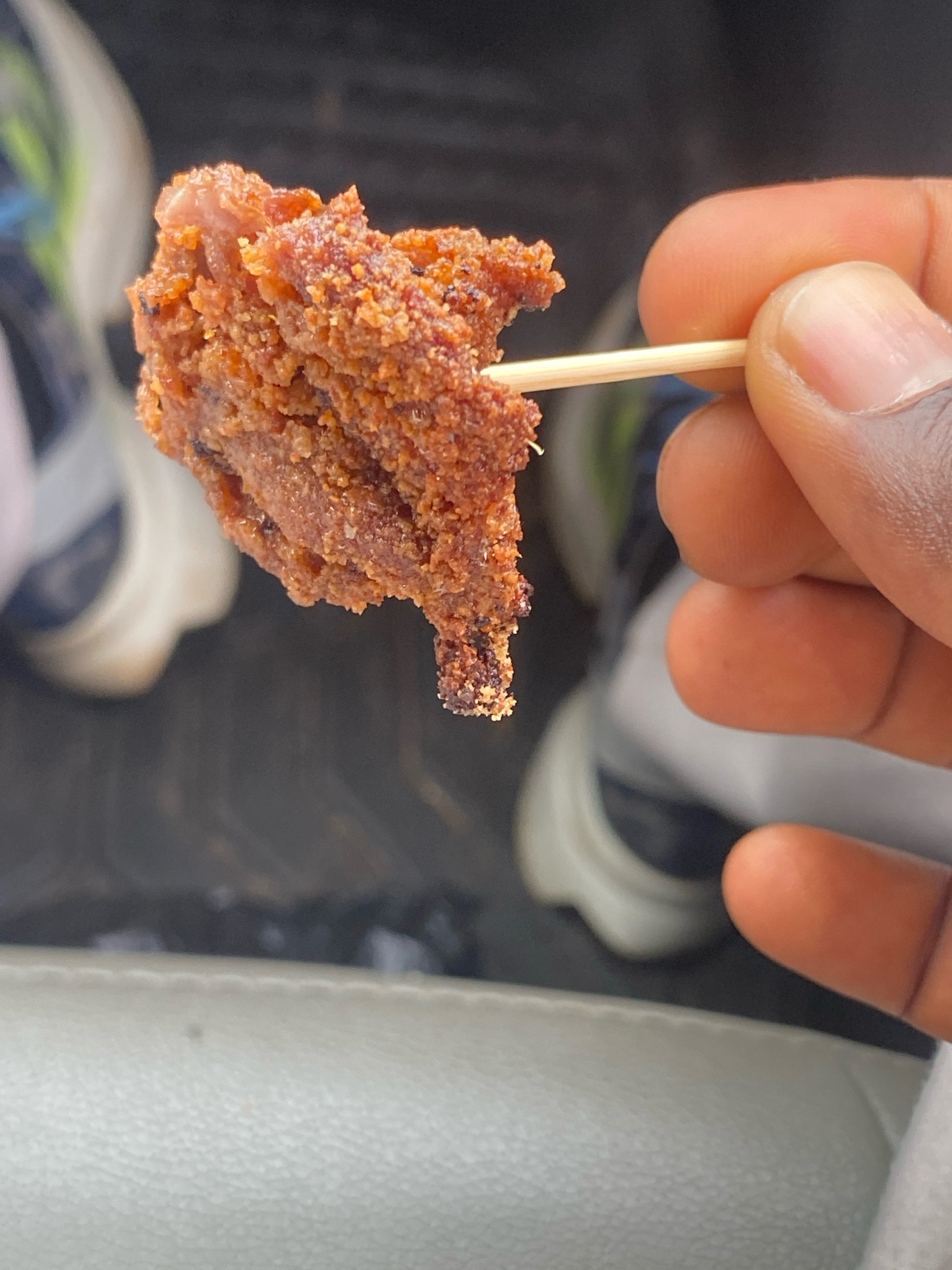 A close-up shot shows a person's hand holding a toothpick with a piece of fried meat. The meat is brown and appears to be coated in spices. The background is out of focus, but suggests an outdoor setting with what look like shoes and a vehicle interior. The image was taken in Kaduna, Nigeria.