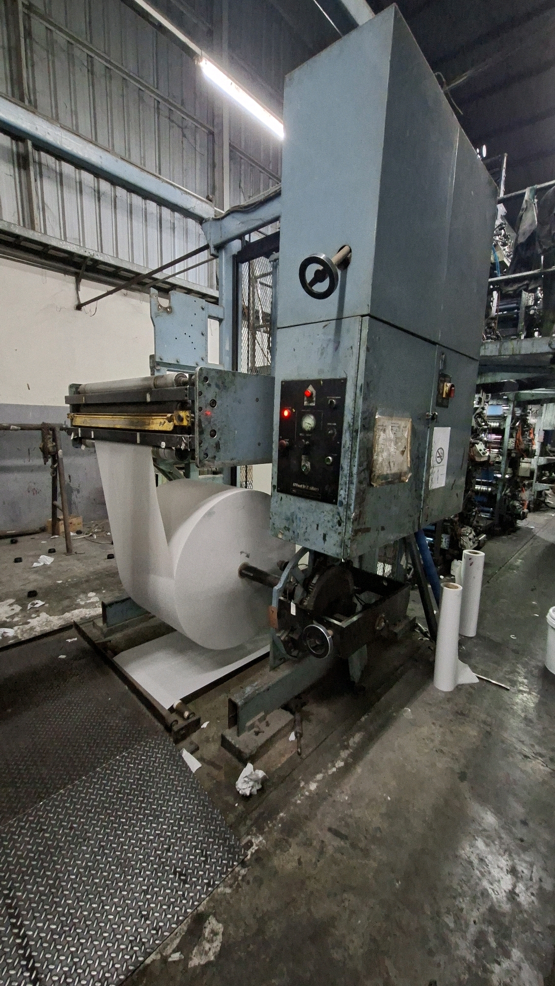 The image shows a large, industrial paper roll feeding machine in what appears to be a dimly lit factory or warehouse. The machine is made of metal, predominantly painted a faded blue-grey. A large roll of white paper is mounted on a spindle, with the paper feeding into the machine. There are control panels with lights and gauges on the side of the machine. The floor is concrete and appears dirty, with some scattered debris. In the background, more industrial equipment and shelving units can be seen, suggesting a manufacturing or printing facility. The lighting is primarily from fluorescent fixtures on the ceiling. The overall impression is one of an active, albeit perhaps aging, industrial workspace. There are no people visible in the image. The specific location in Palembang, Indonesia, cannot be determined from the visual information provided.